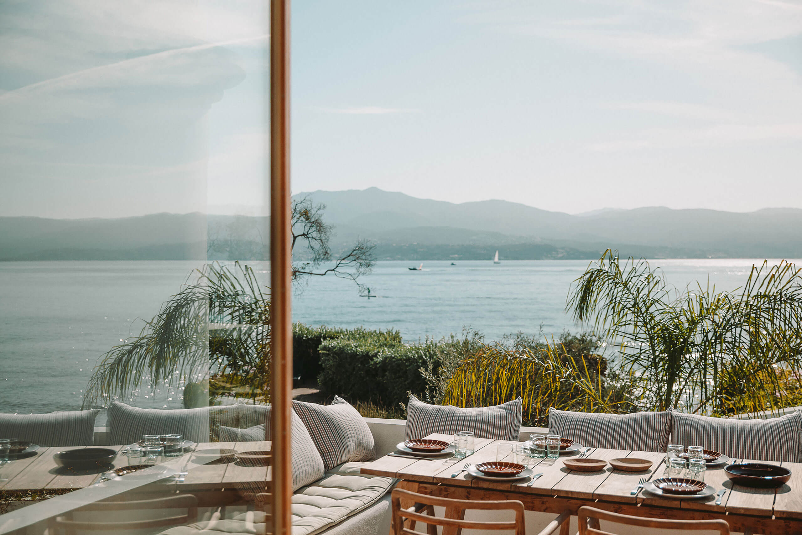 Casa Santa Teresa, Europe — Lakeside dining terrace with striped seating, brass-framed glass doors, ceramic tableware, mountain views, and waterfront garden with native grasses.