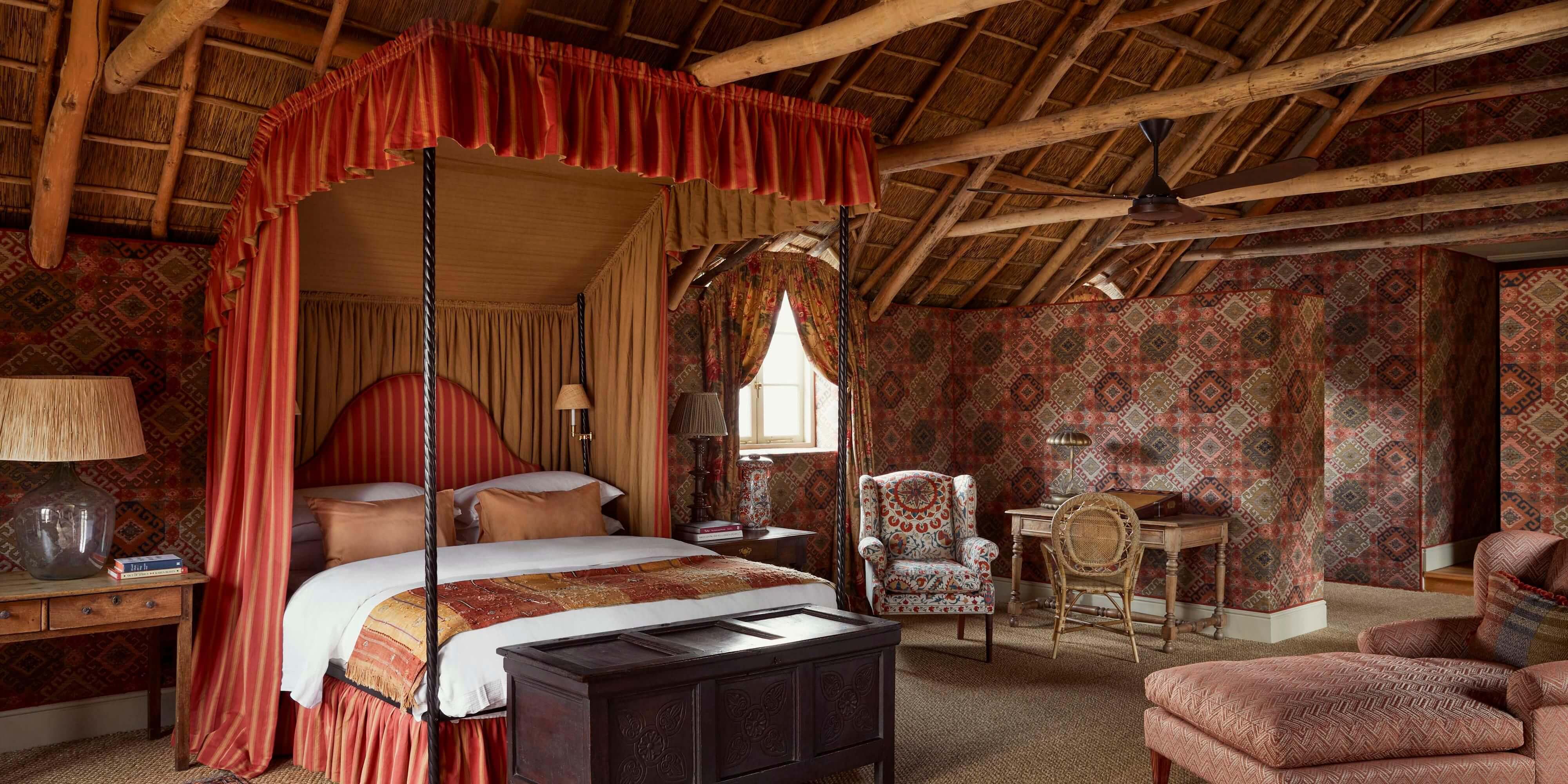 Sterrekopje, Africa — Luxury four-poster bedroom with red silk drapes, patterned wallpaper, thatched ceiling, period furnishings and warm natural light from casement window.