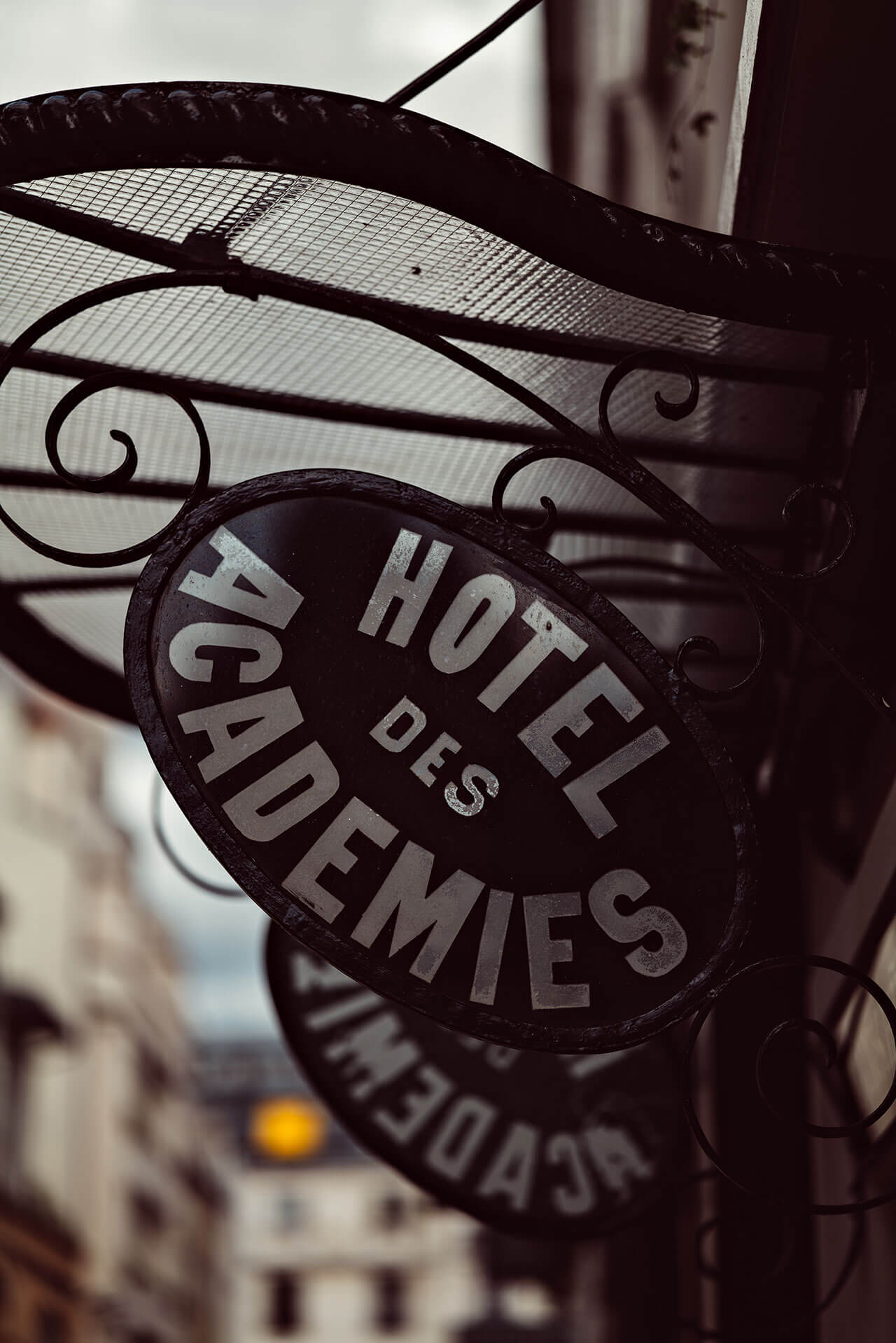 Académies et des Arts, Europe — Ornate black wrought-iron hotel signage with gilt lettering, architectural detail with scrollwork and mesh, soft natural light casting shadows.
