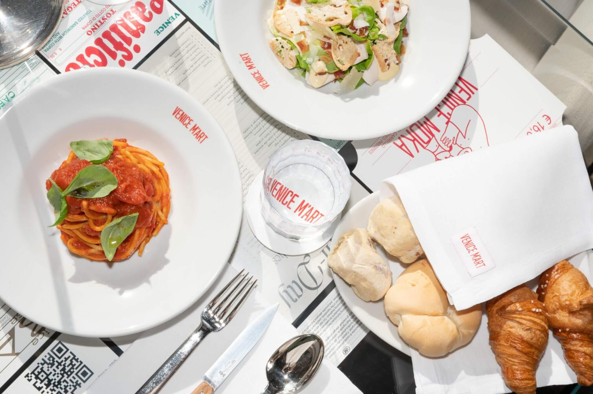Venice M’Art All-day Restaurant — Venice M'Art All-day Restaurant, Europe — Overhead view of Italian pasta with tomato sauce and basil, fresh seafood salad, croissants, and branded tableware in minimalist white dining setting.