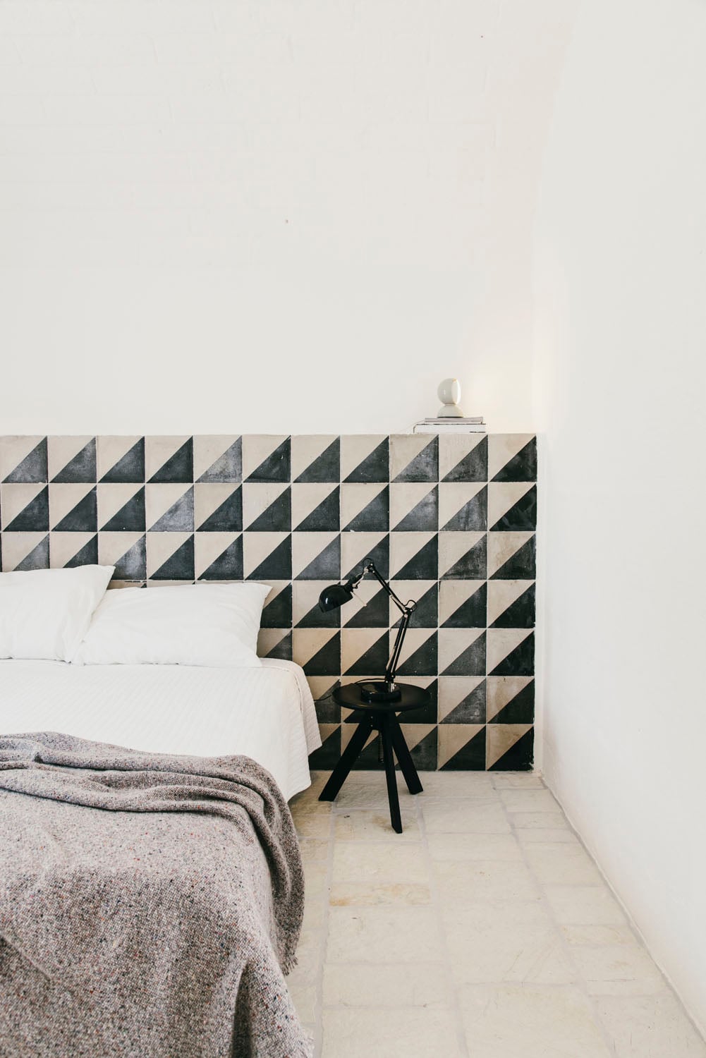 Masseria Moroseta, Europe — Minimalist bedroom with geometric black and white tile headboard, white linens, gray throw blanket, black tripod lamp, pale stone floor, and stark white walls.