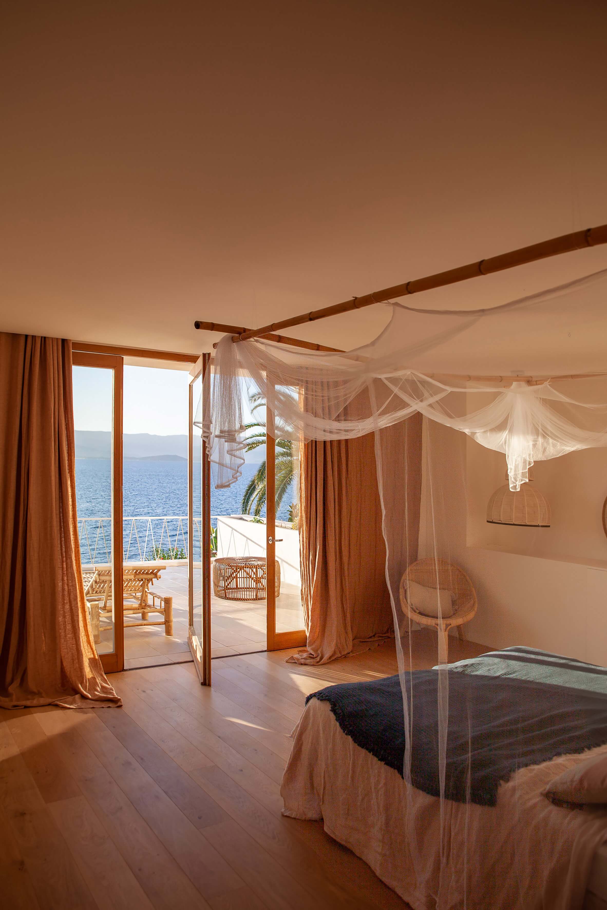 Casa Santa Teresa, Europe — Seafront bedroom with sheer canopy bed, wooden beams, ochre curtains, and open terrace overlooking Mediterranean waters at golden hour.