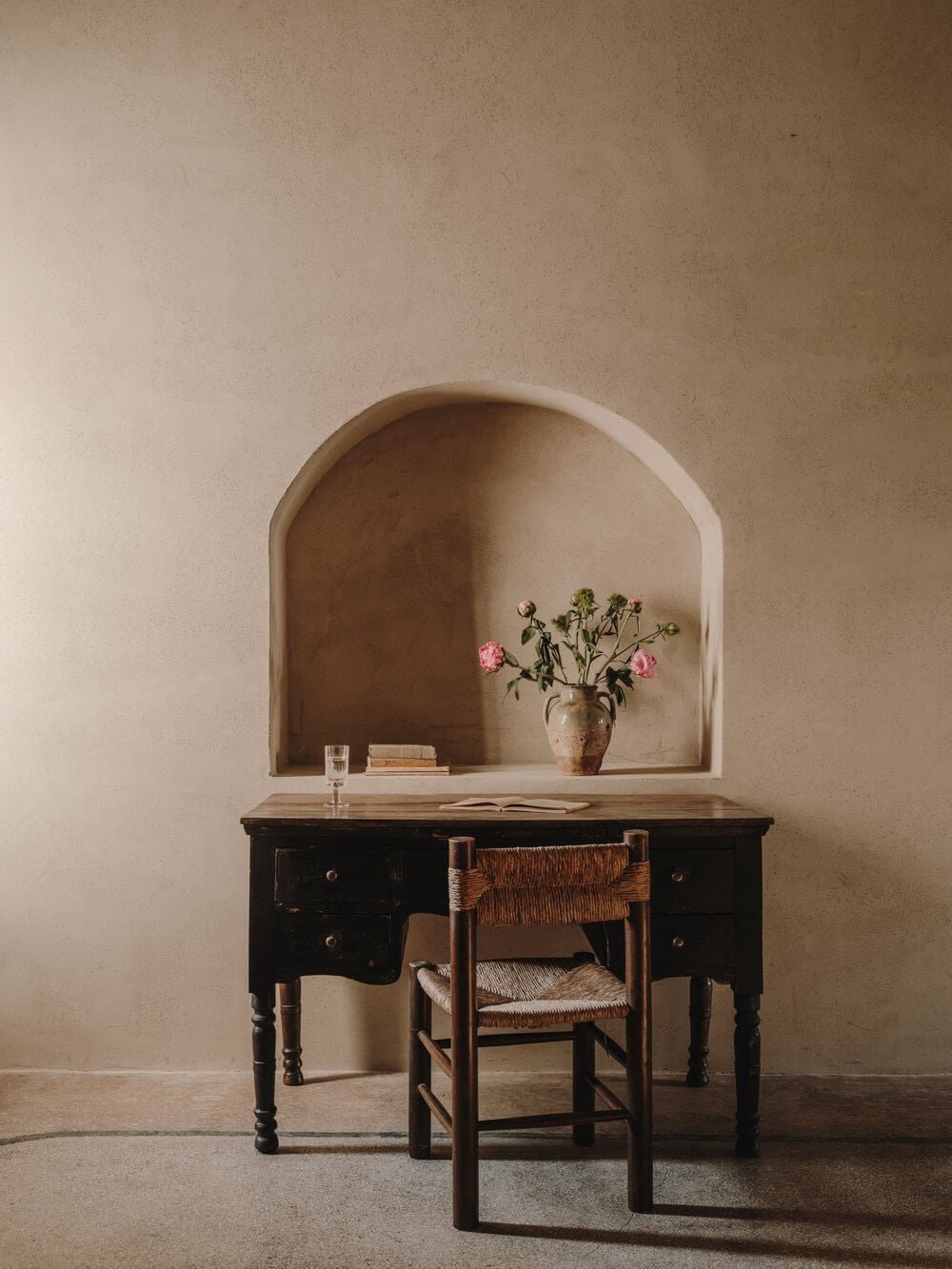 Casa Soleto, Europe — Minimalist desk with woven chair beneath arched alcove, pink flowers in terracotta vase, warm plaster walls with natural light.