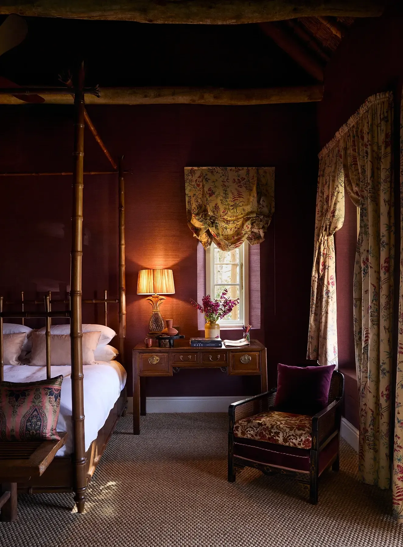 Sterrekopje, Africa — Candlelit bedroom with bamboo four-poster bed, burgundy walls, floral curtains, antique desk, and warm ambient lighting.