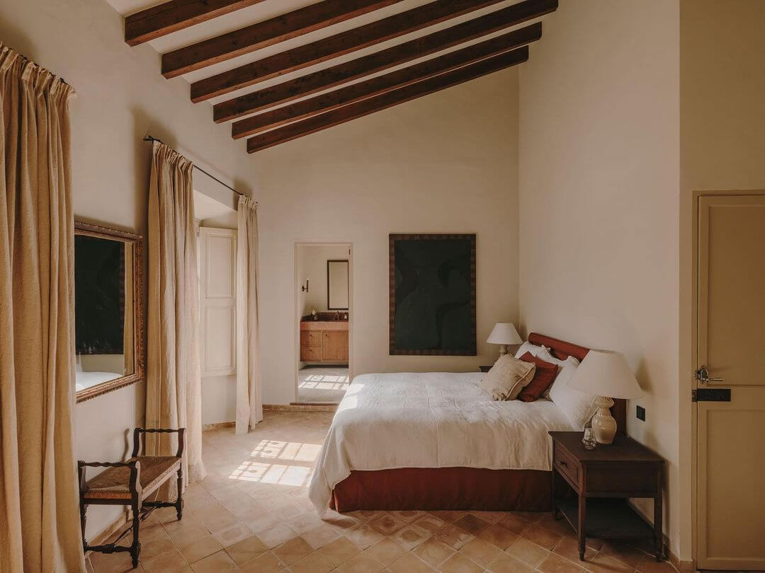 Santa Teresa 34, Balearic Islands, Spain — Sunlit bedroom with exposed wooden beams, terracotta flooring, neutral linens, and warm earthy tones.
