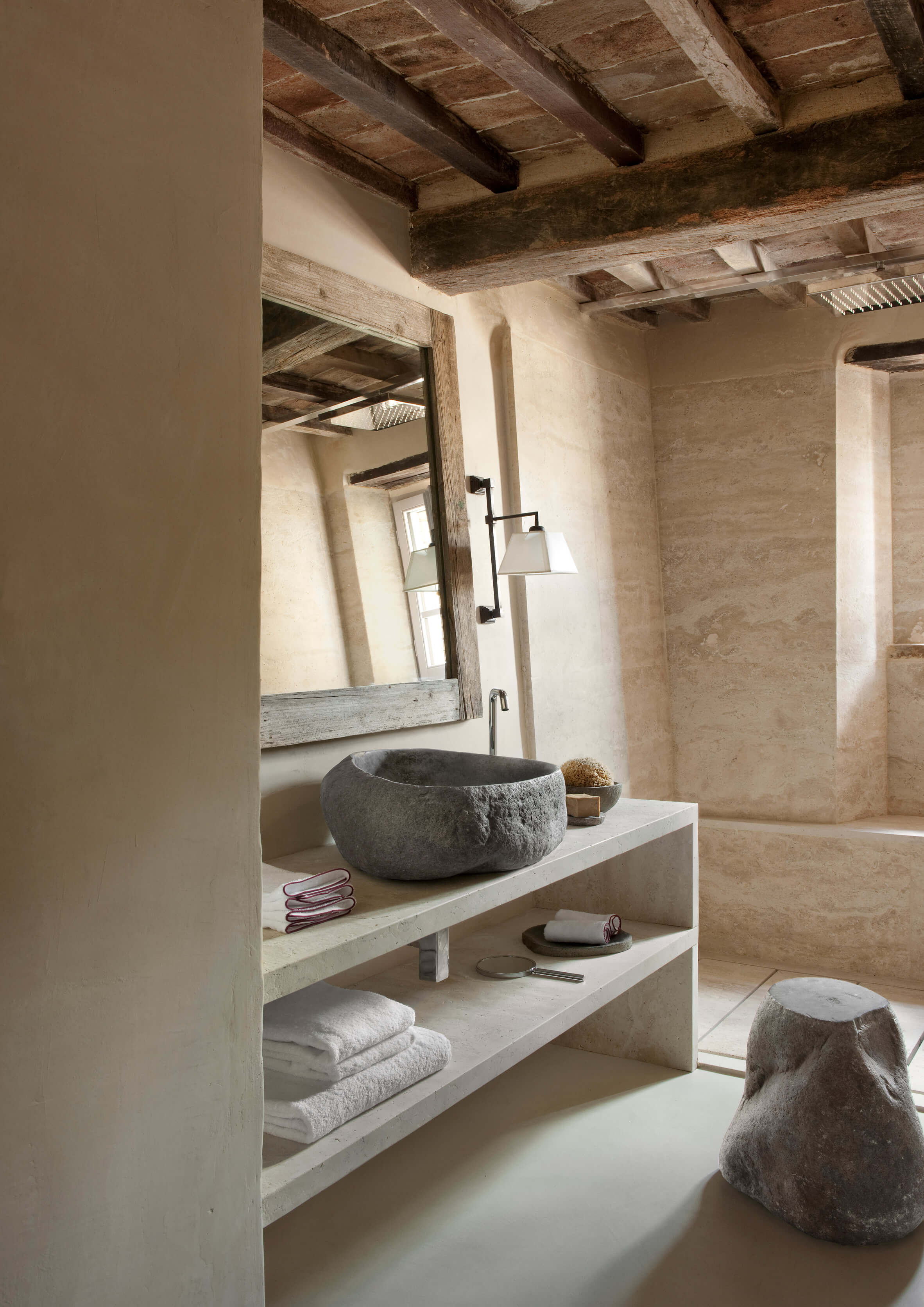 Monteverdi, Europe — Minimalist bathroom with natural stone vessel sink, wooden beam ceiling, plaster walls, and open shelving with linens and toiletries.
