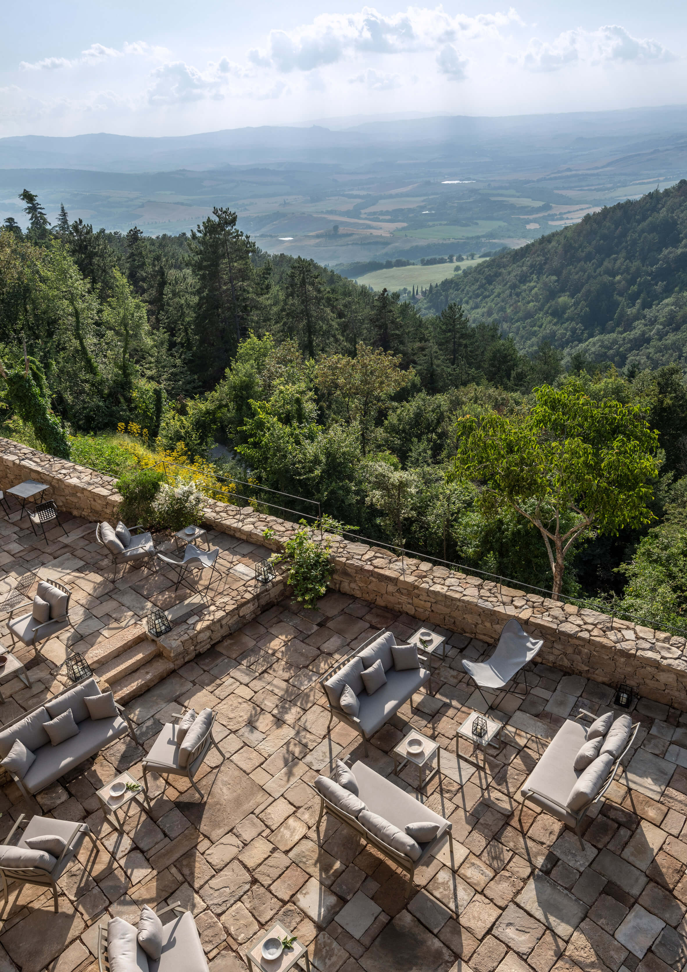 Monteverdi, Europe — Travertine terrace with contemporary seating overlooks forested valley and distant Tuscan hills under soft afternoon light.
