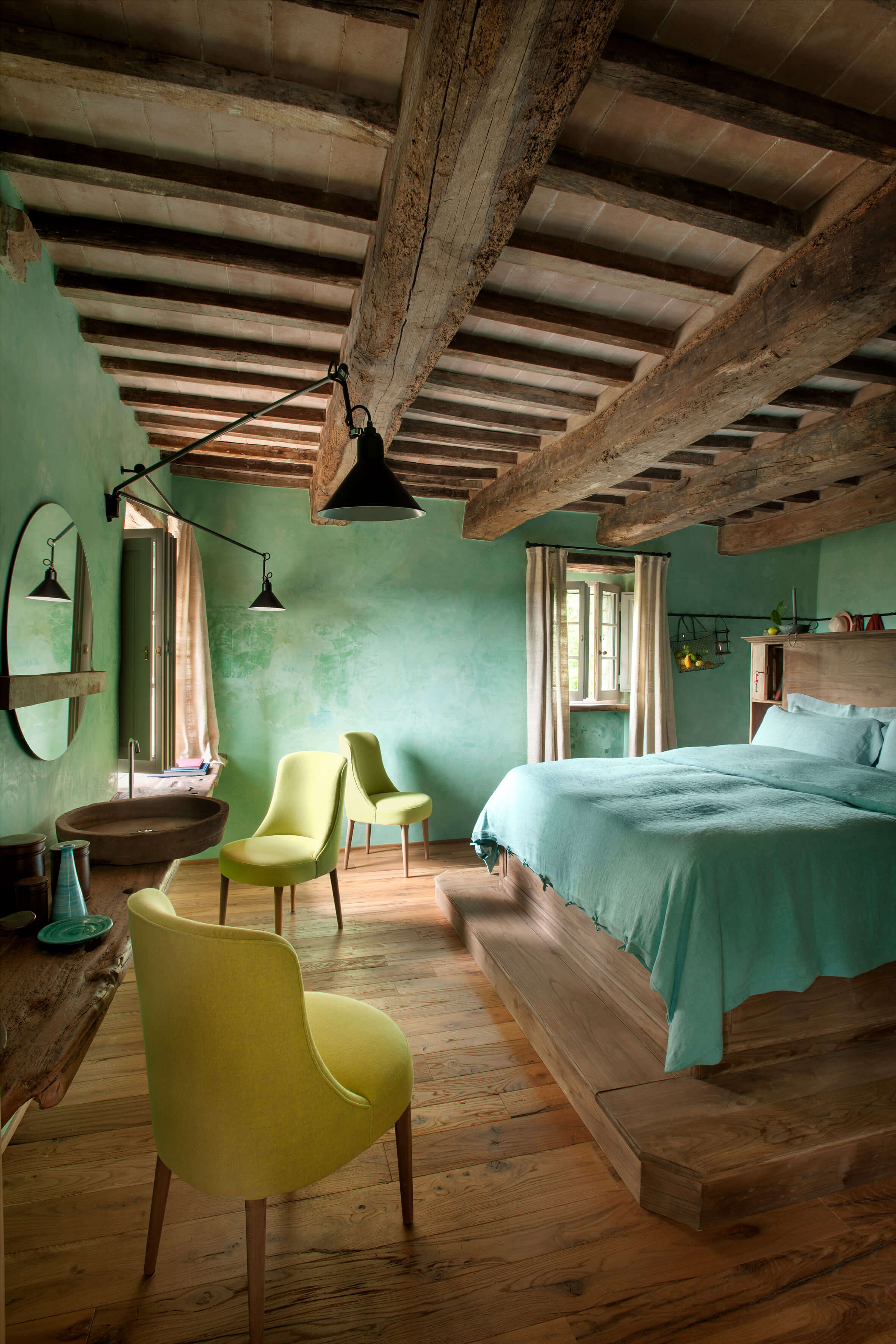 Monteverdi, Europe — Bedroom with exposed timber beams, green plaster walls, pale yellow chairs, turquoise bedding, wooden floors, and black pendant lights.