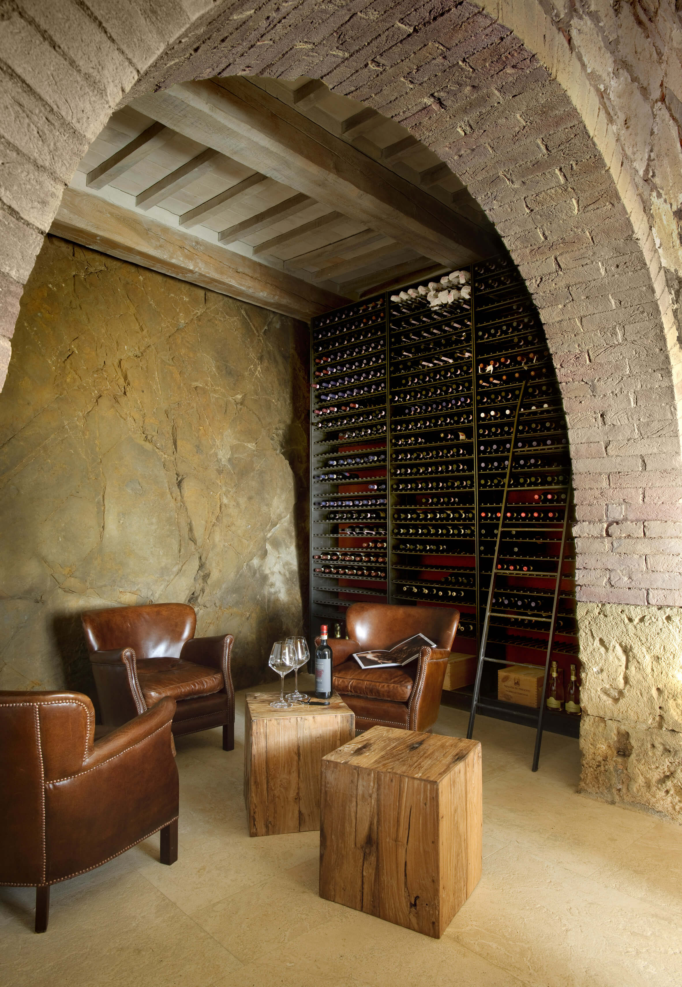 Monteverdi, Europe — Arched stone wine cellar with leather club chairs, wooden side tables, and floor-to-ceiling wine racks under wooden beams.