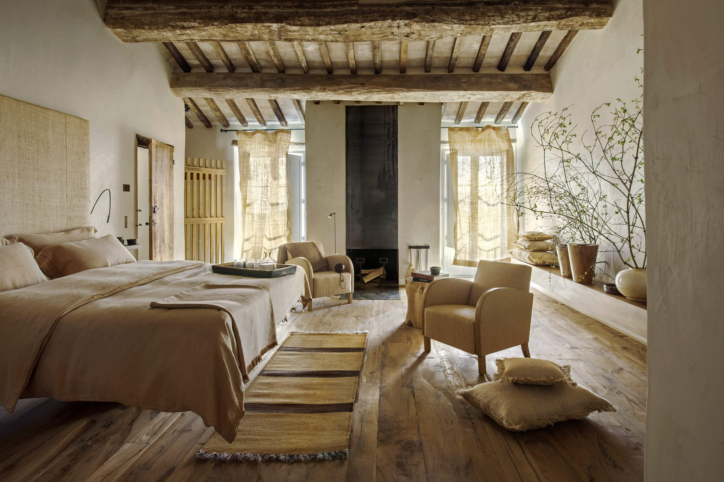 Monteverdi, Europe — Rustic bedroom with exposed wooden beams, neutral linens, fireplace, natural light through sheer curtains, terracotta floors, modern minimalist furnishings.