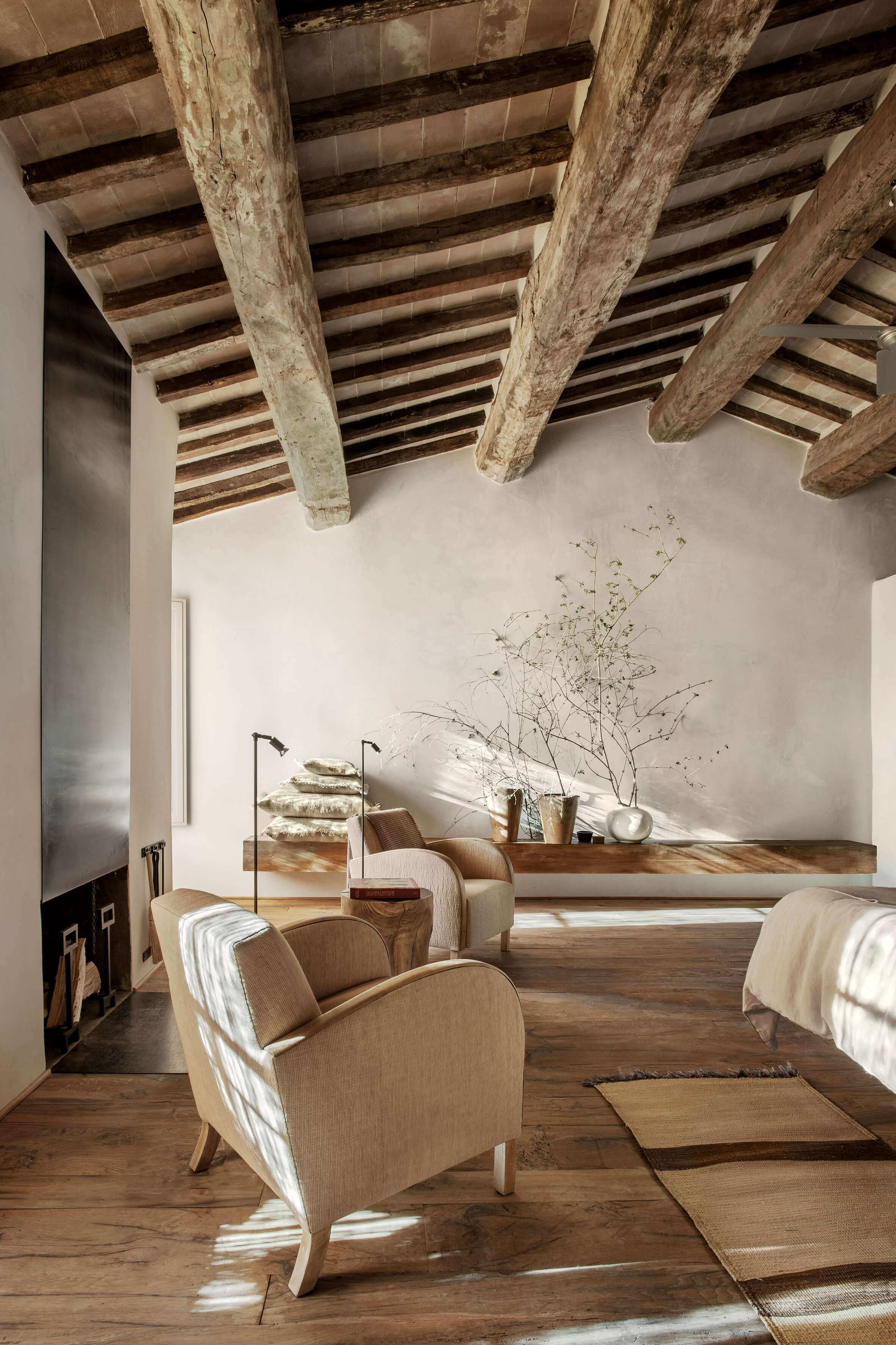 Monteverdi, Europe — Rustic bedroom with exposed wooden beams, neutral linen armchair, wooden shelf with dried botanicals, warm natural light streaming across wooden floors.