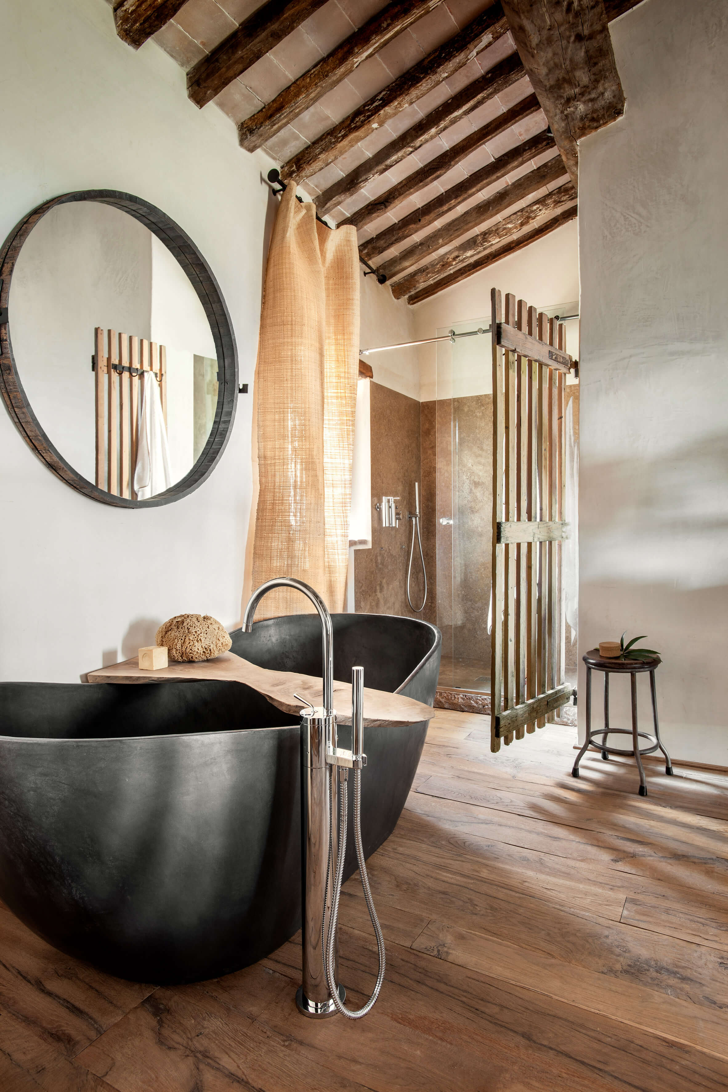 Monteverdi, Europe — Black soloetta bathtub with wooden tray, round mirror, exposed beam ceiling, and walk-in shower with natural light in luxury spa bathroom.