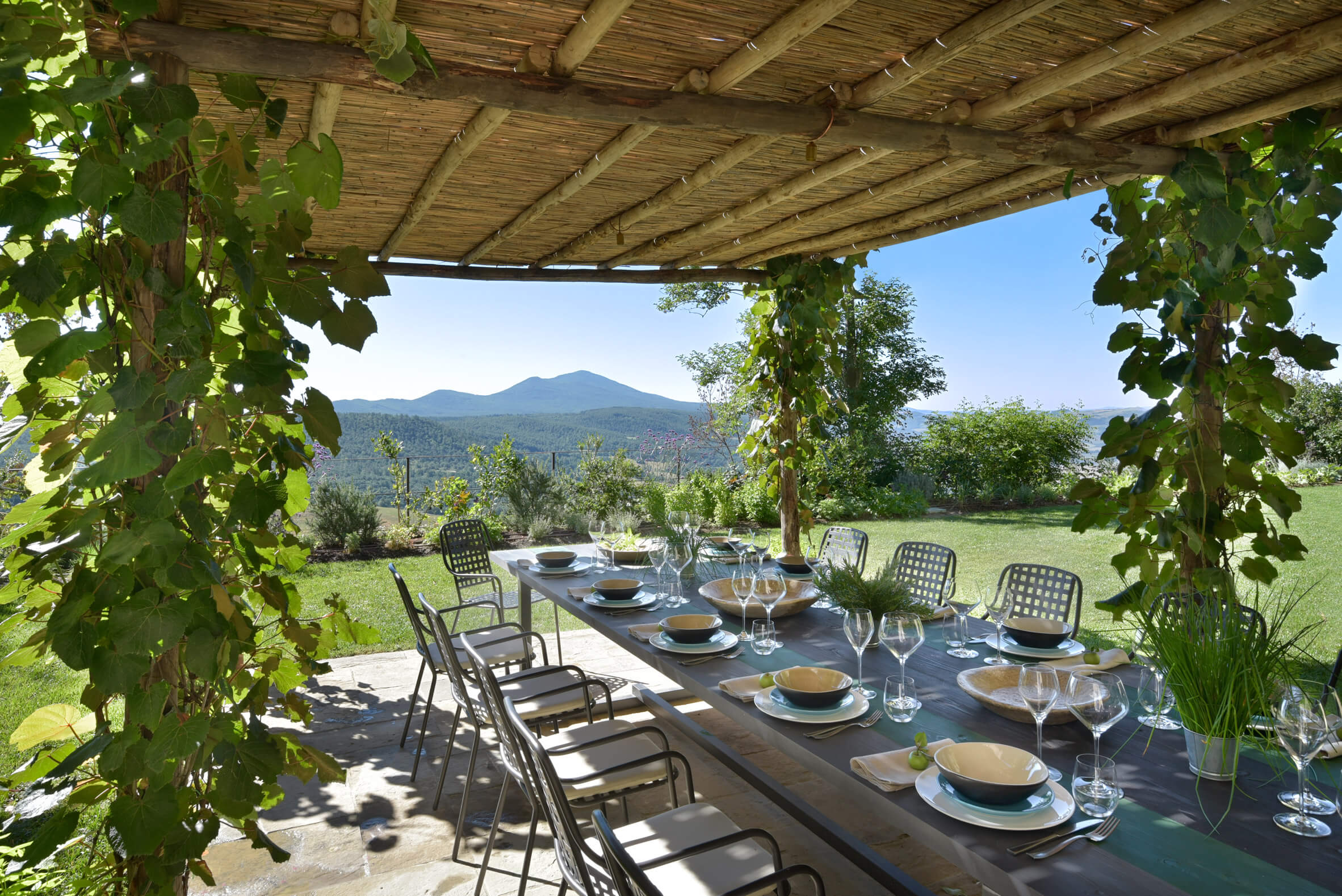 Monteverdi, Europe — Long dining table set beneath wooden pergola with grapevines, mountain views, manicured gardens, natural light.