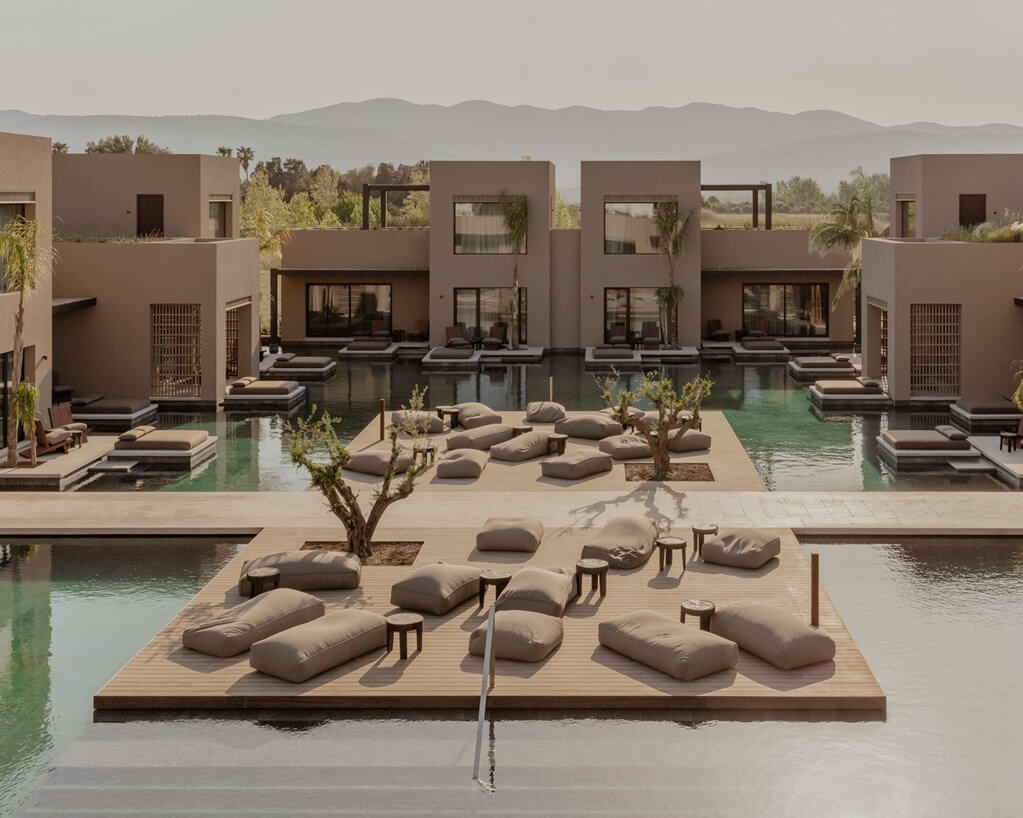 Casa Cook Samos, Greece — Minimalist clay-colored villas with turquoise pools, floating wooden platforms with linen cushions, palm trees and mountains beyond.