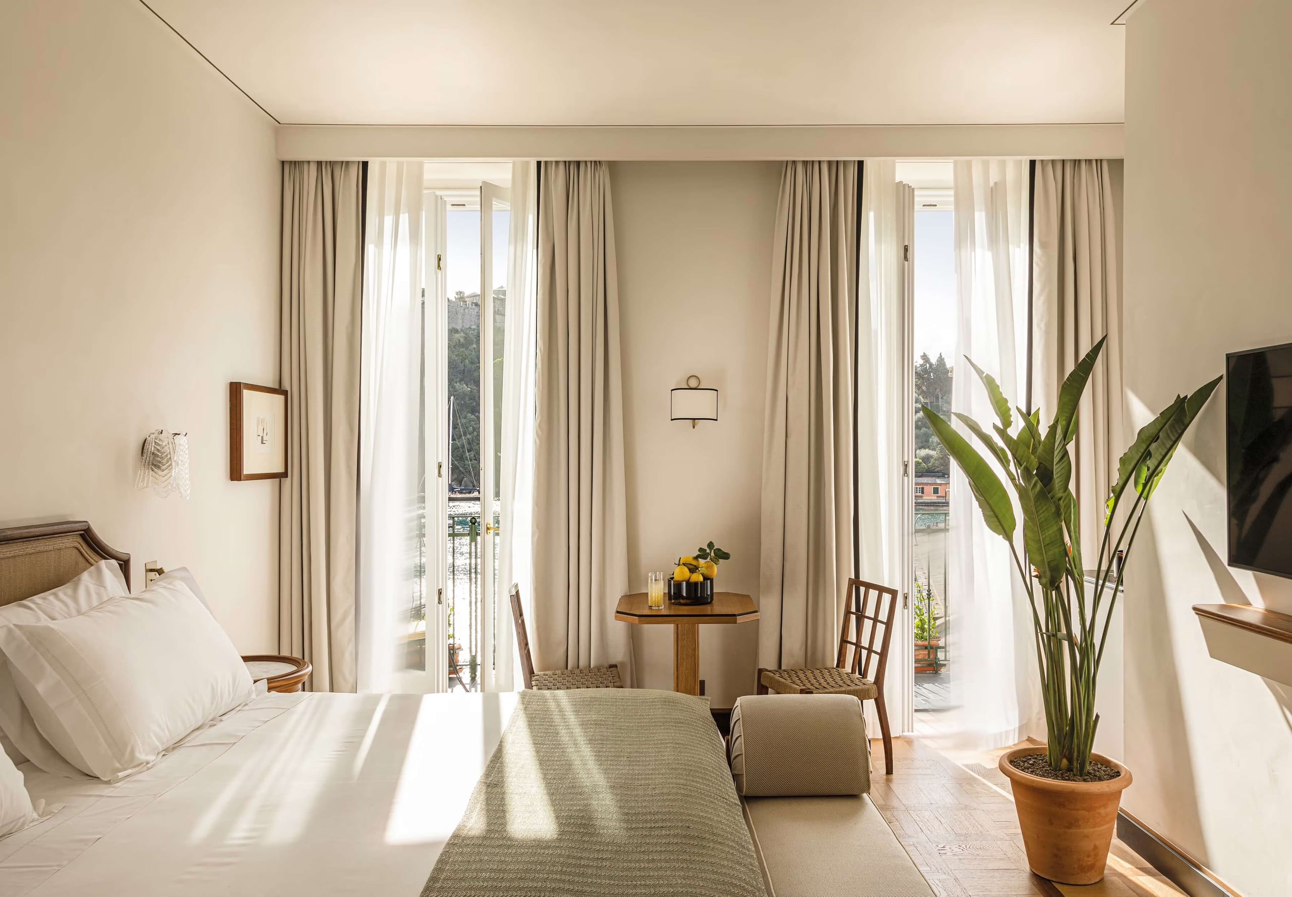 Splendido Mare, Europe — Sunlit bedroom suite with cream linens, wooden furniture, floor-to-ceiling windows, sheer curtains, potted plant, and coastal views.