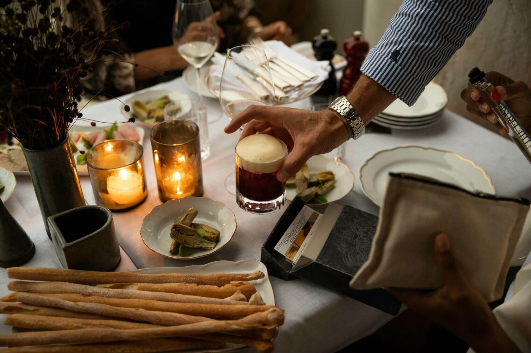 Pre-Dinner At The Venice Venice Hotel — Pre-Dinner At The Venice, Venice, Europe — candlelit fine dining table with craft cocktail, artisanal breadsticks, plated appetizers, and minimalist tableware in luxury hospitality setting.