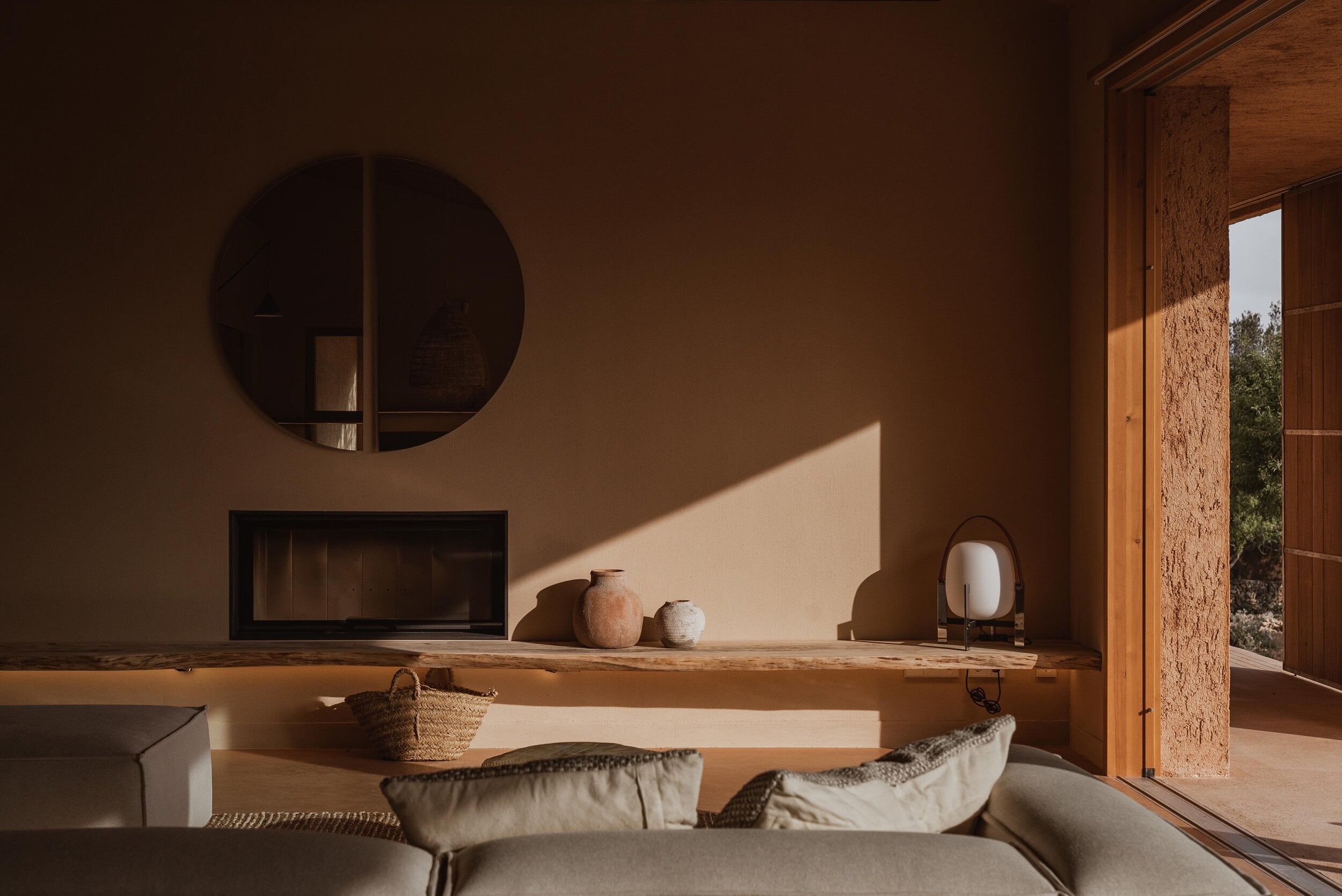 Alcúdia House, Balearic Islands — Minimalist bedroom with terracotta walls, circular mirror, fireplace, wooden mantelpiece, natural light, cork architectural details, and open doors to landscape.