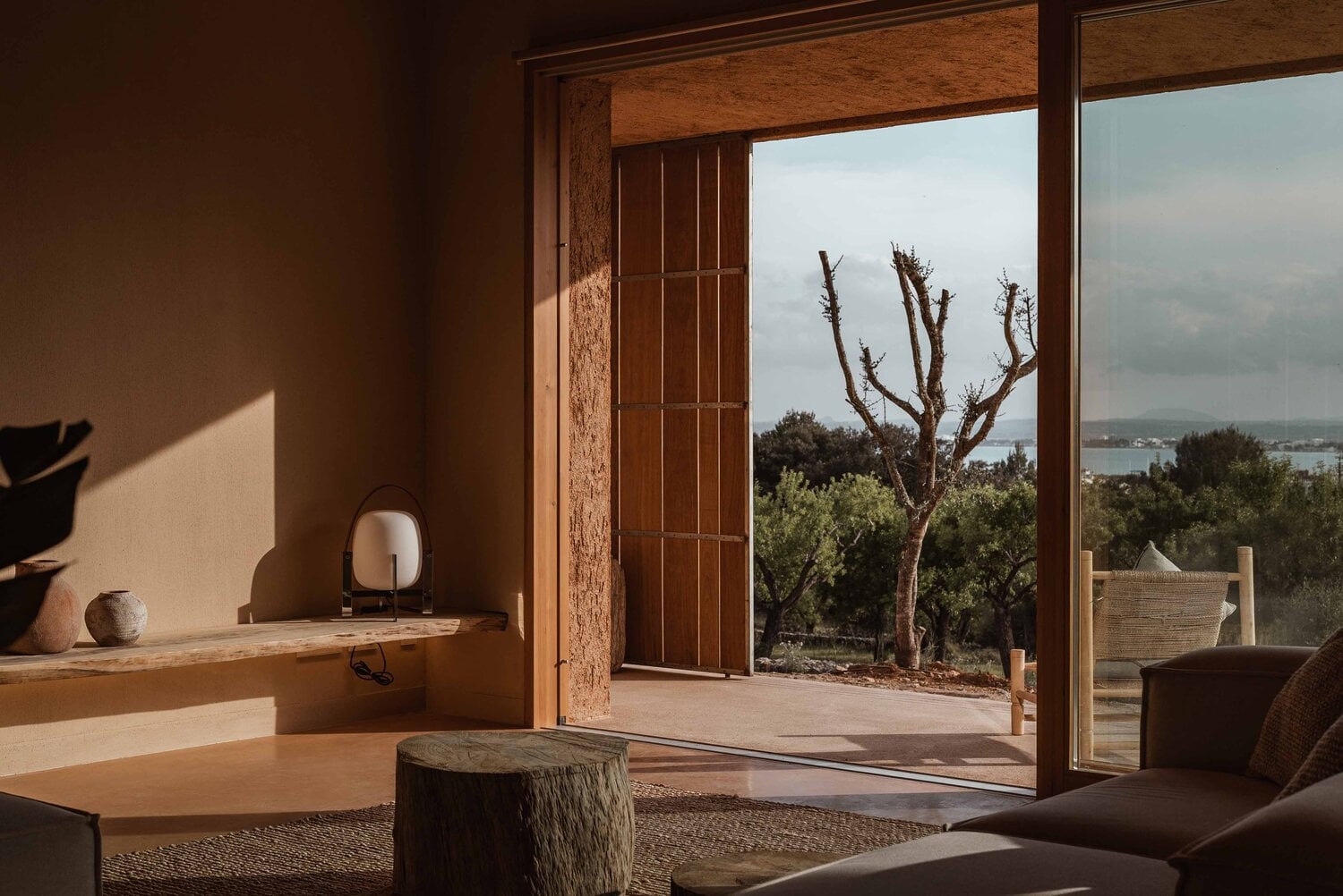 Alcúdia House, Balearic Islands, Spain — Minimalist interior with cork walls, wooden beams, and open terrace overlooking olive groves and Mediterranean sea.