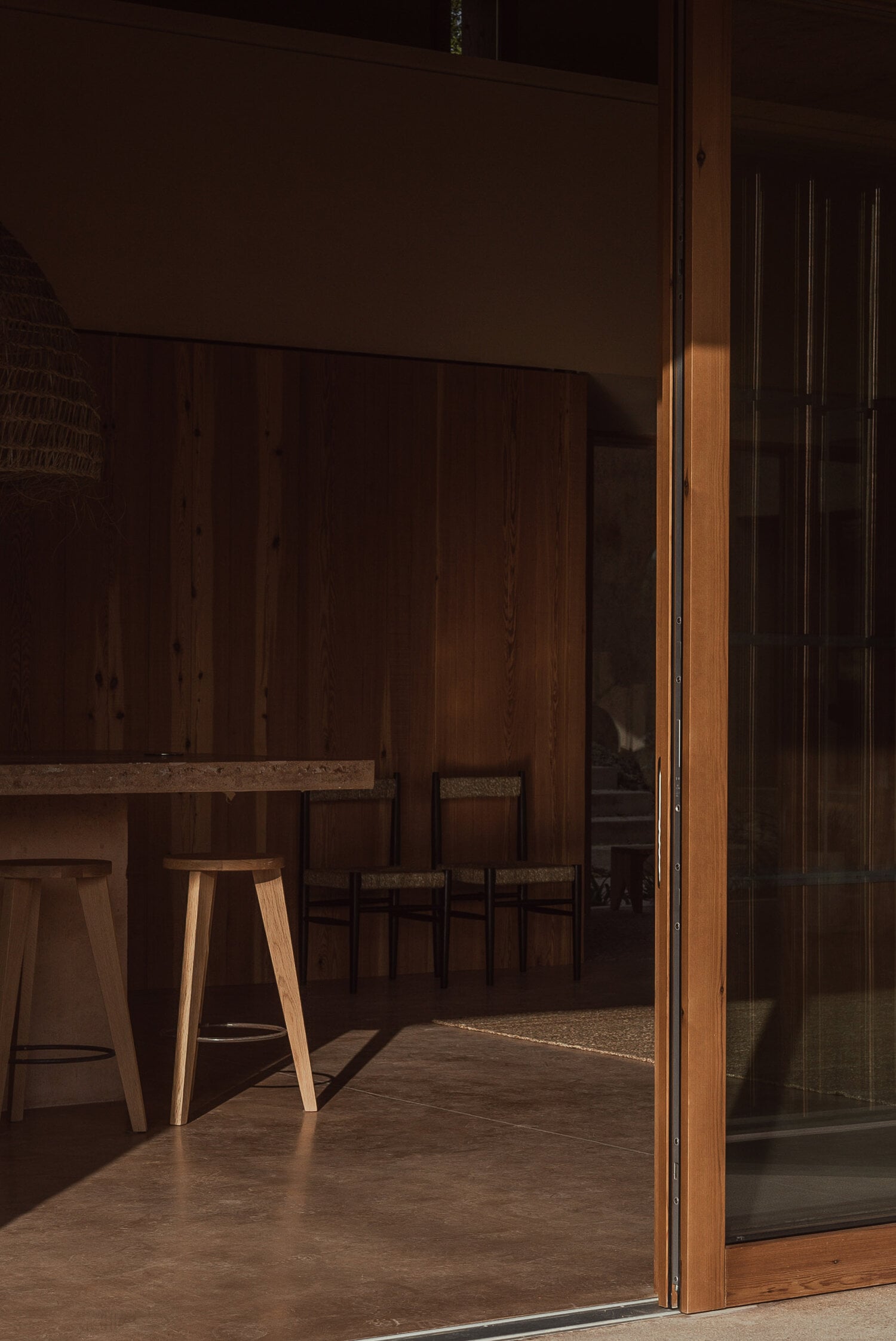 Alcúdia House, Balearic Islands — Minimalist wooden interior with natural light casting shadows across concrete floors, lounge seating visible through open glass doors.