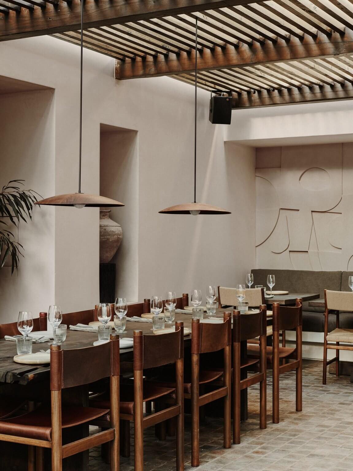 Nōema, Europe — Minimalist dining room with wooden chairs, pendant lighting, striped timber pergola, and sculptural wall relief in neutral palette.