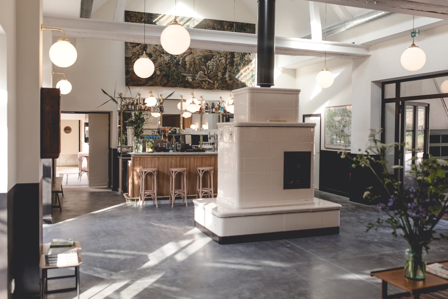 Le Barn, Europe — Contemporary hotel lobby with polished concrete floors, sculptural white fireplace, globe pendant lights, and natural skylight illuminating minimalist bar and seating areas.