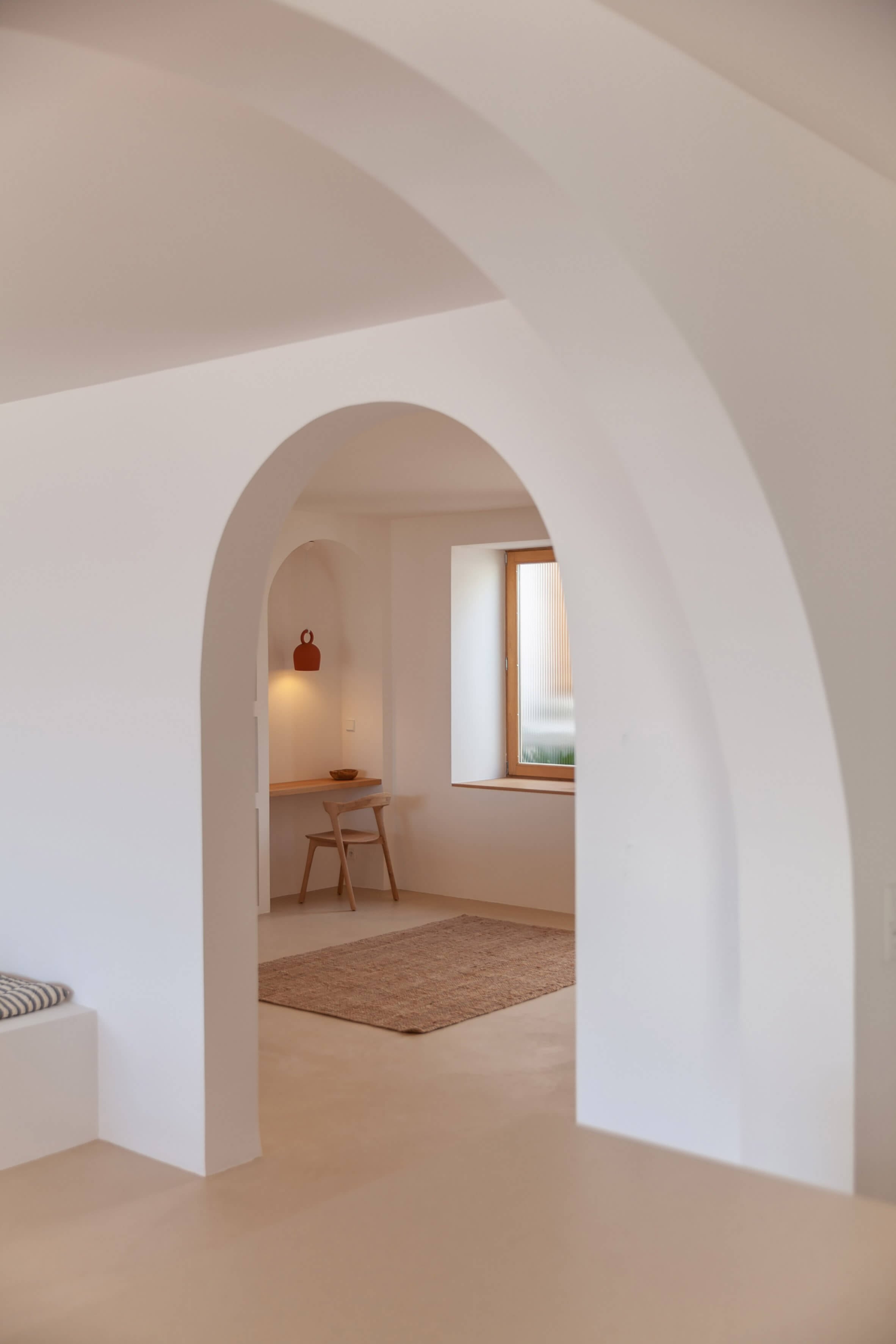 Casa Santa Teresa, Europe — Arched white corridor with natural light, wooden chair, terracotta rug, minimalist Mediterranean architecture.