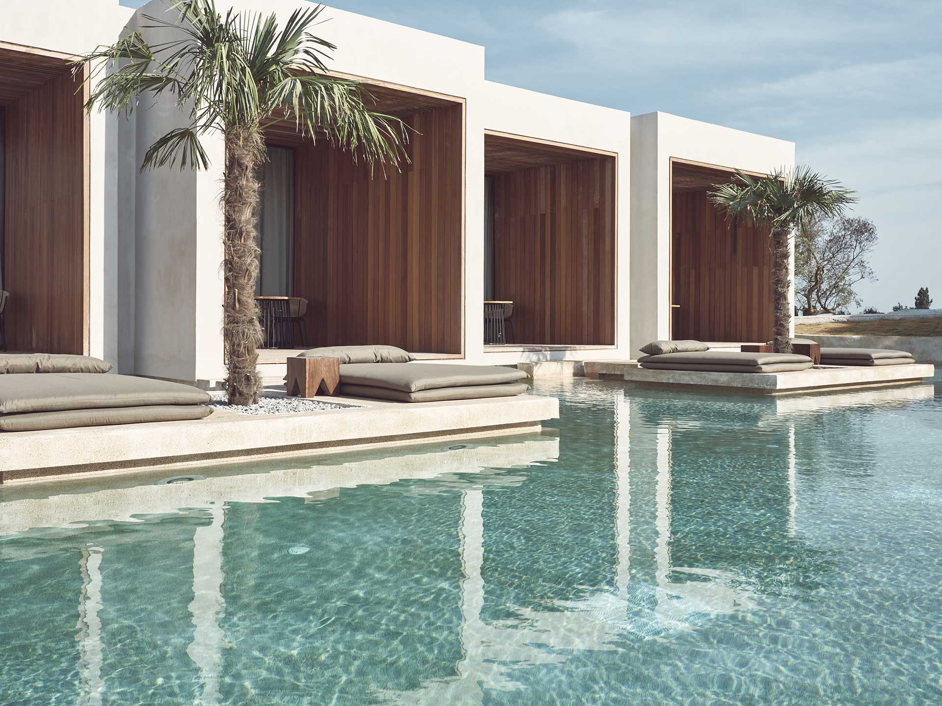 Olea All Suite Hotel, Europe — Minimalist poolside pavilions with dark wood screens, cream loungers, palm trees, and crystal water reflecting white architecture under blue sky.