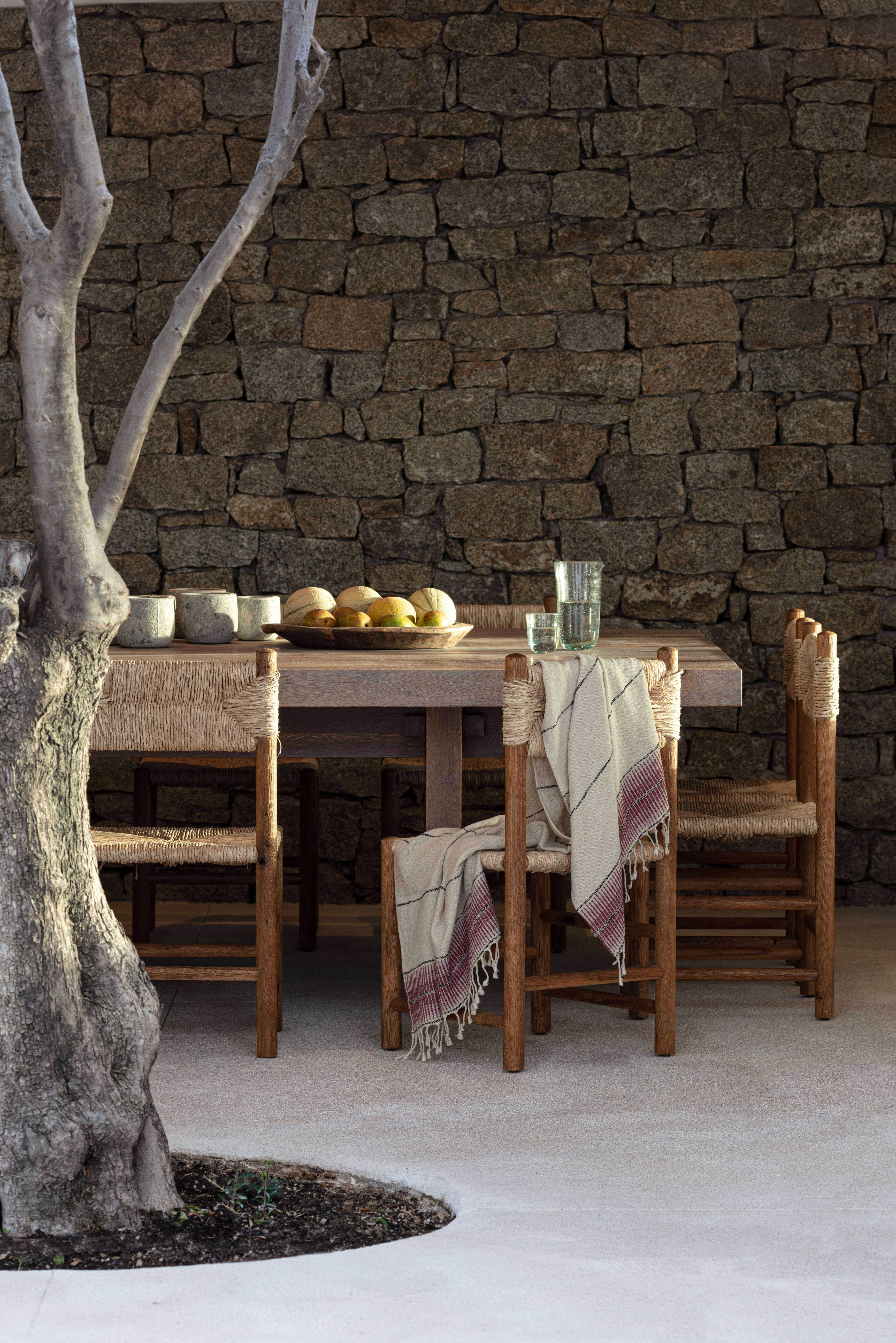 Villa Tama, Europe — Dining table with woven chairs beneath stone wall, afternoon light filtering through olive tree, glasses and fruit bowl on linen-draped table.