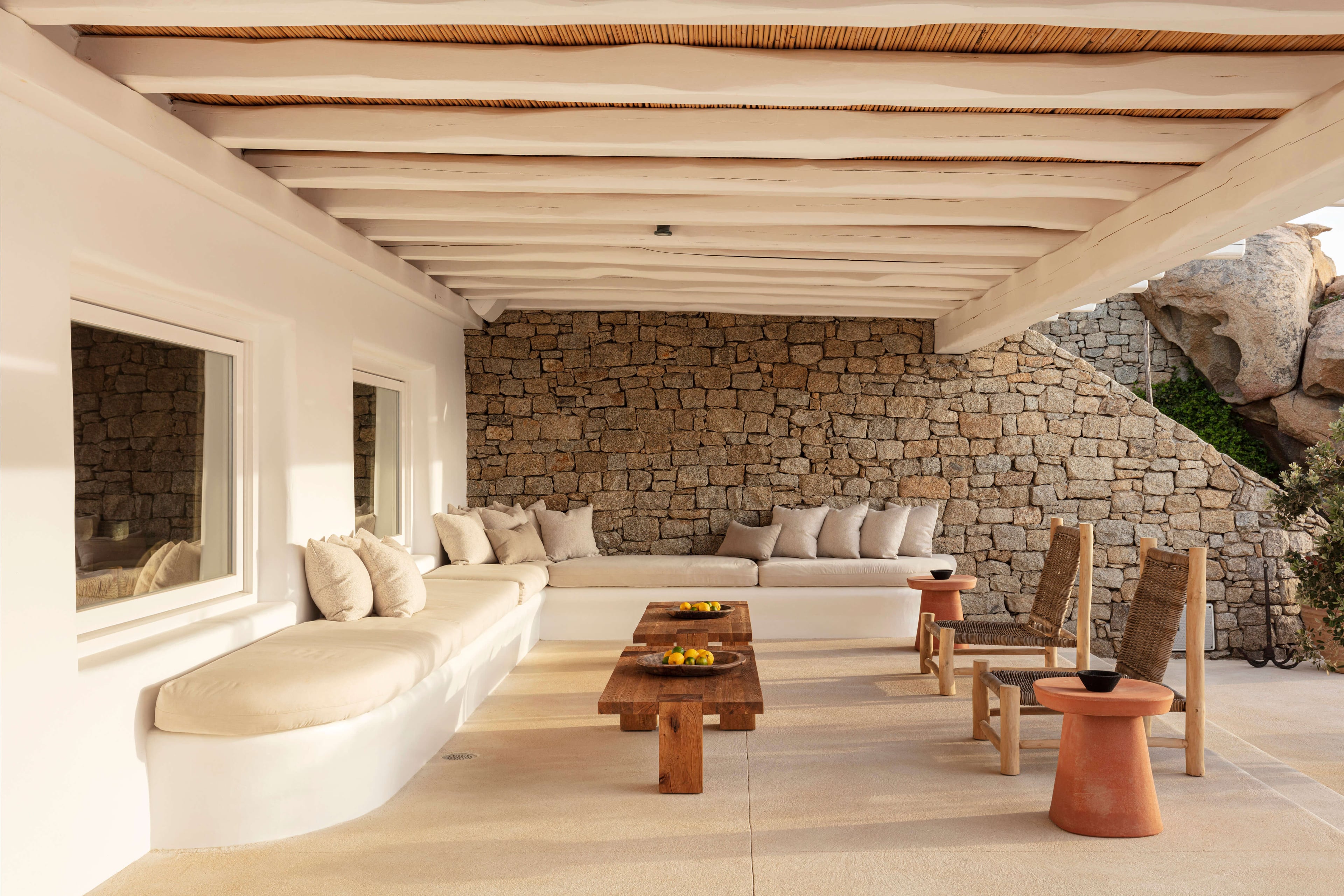 Villa Tama, Europe — Covered terrace with curved cream seating, stone wall, wooden beams, natural light, woven chairs, and terracotta accents.
