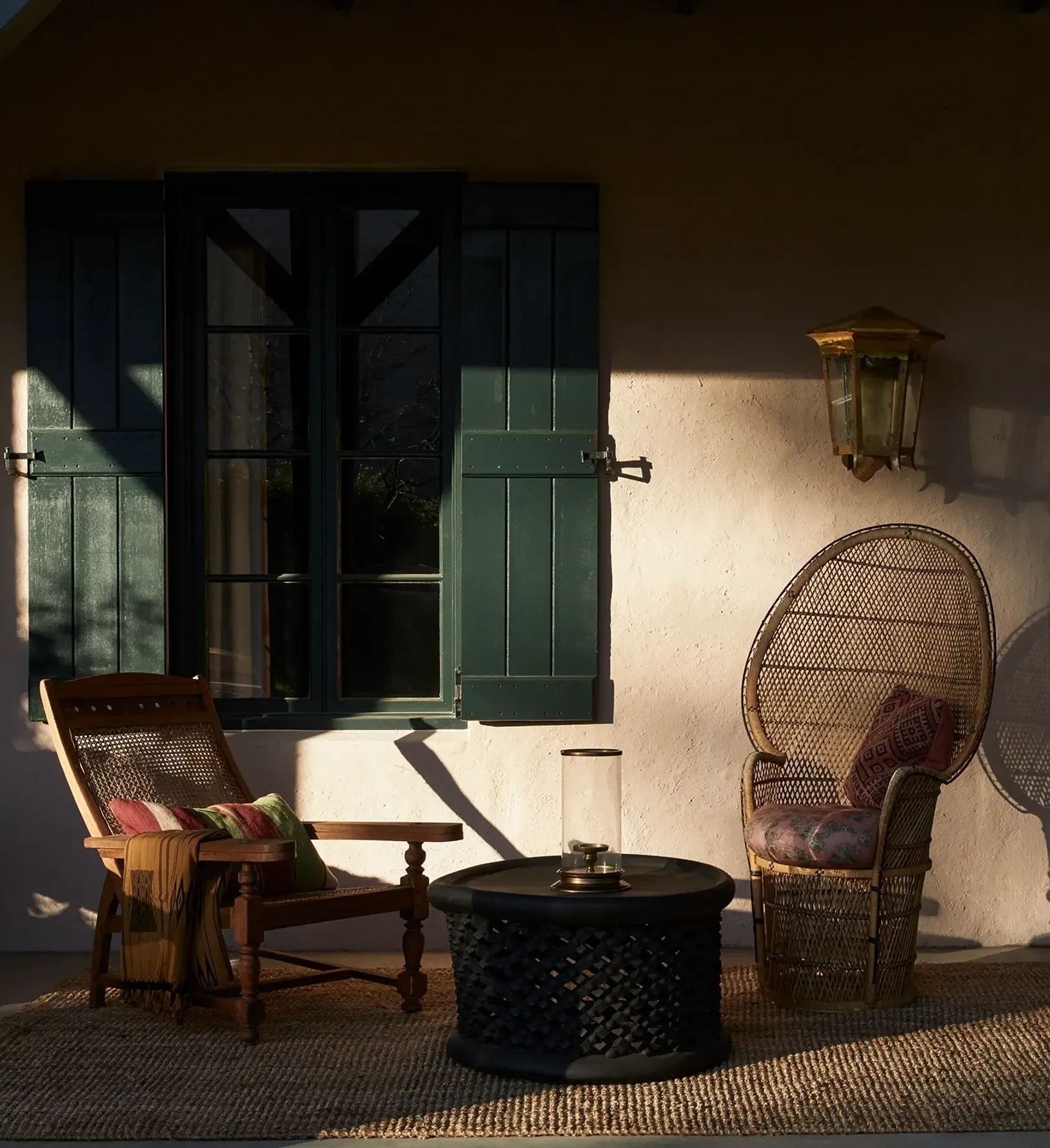 Sterrekopje, Africa — Shaded courtyard with woven rattan chairs, black side table, cream stucco walls, and green shuttered doors casting geometric shadows in golden light.