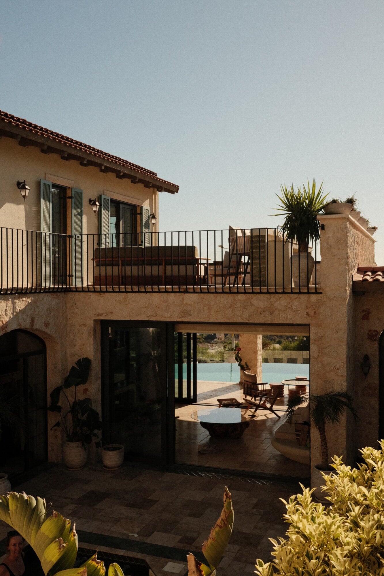Villa Thalia by Muses Kalkan, Kalkan, Turkey — Ochre stone villa with black-framed glass doors opening to pool terrace, upper-level balcony with iron railing, palm plants, clear Mediterranean light.