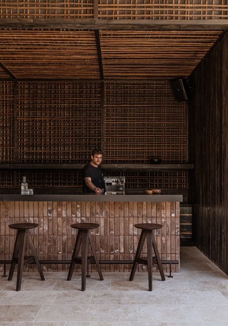 Parea Bar, Europe — Minimalist brick bar interior with wooden counter, dark timber stools, and geometric wall detailing under linear ceiling elements.