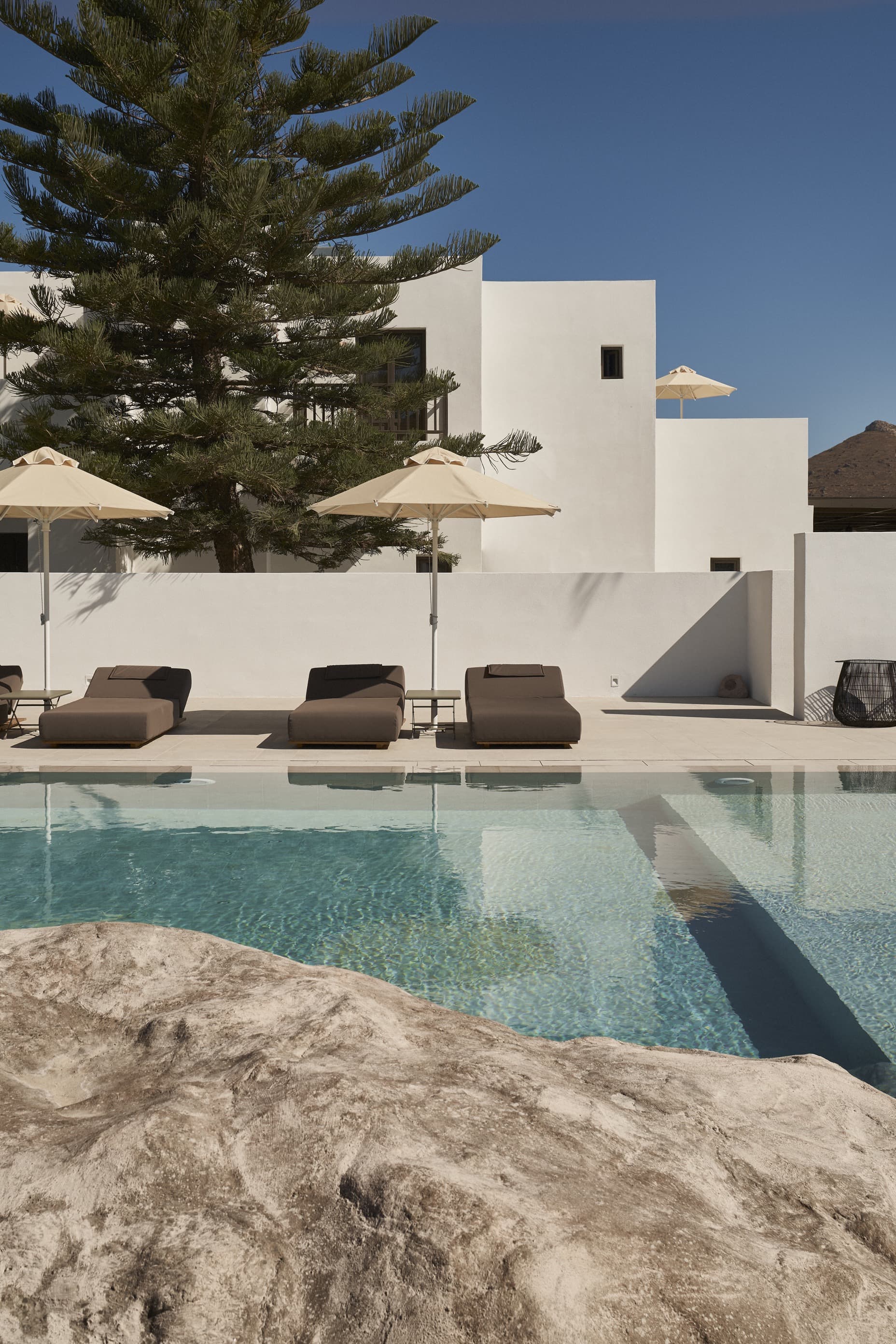 Parīlio Hotel, Europe — Minimalist white architecture with infinity pool, taupe loungers, striped umbrellas, and Mediterranean pine tree against clear blue sky.