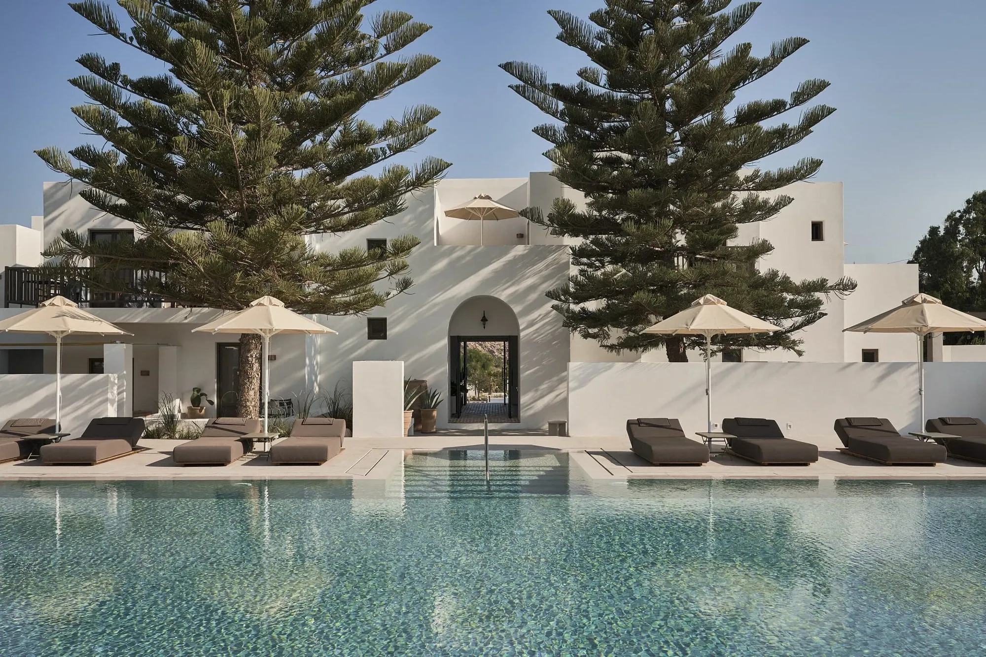 Parīlio Hotel, Europe — whitewashed Mediterranean resort with arched entryway, infinity pool, loungers, cypress trees, and minimalist courtyard architecture under clear sky.