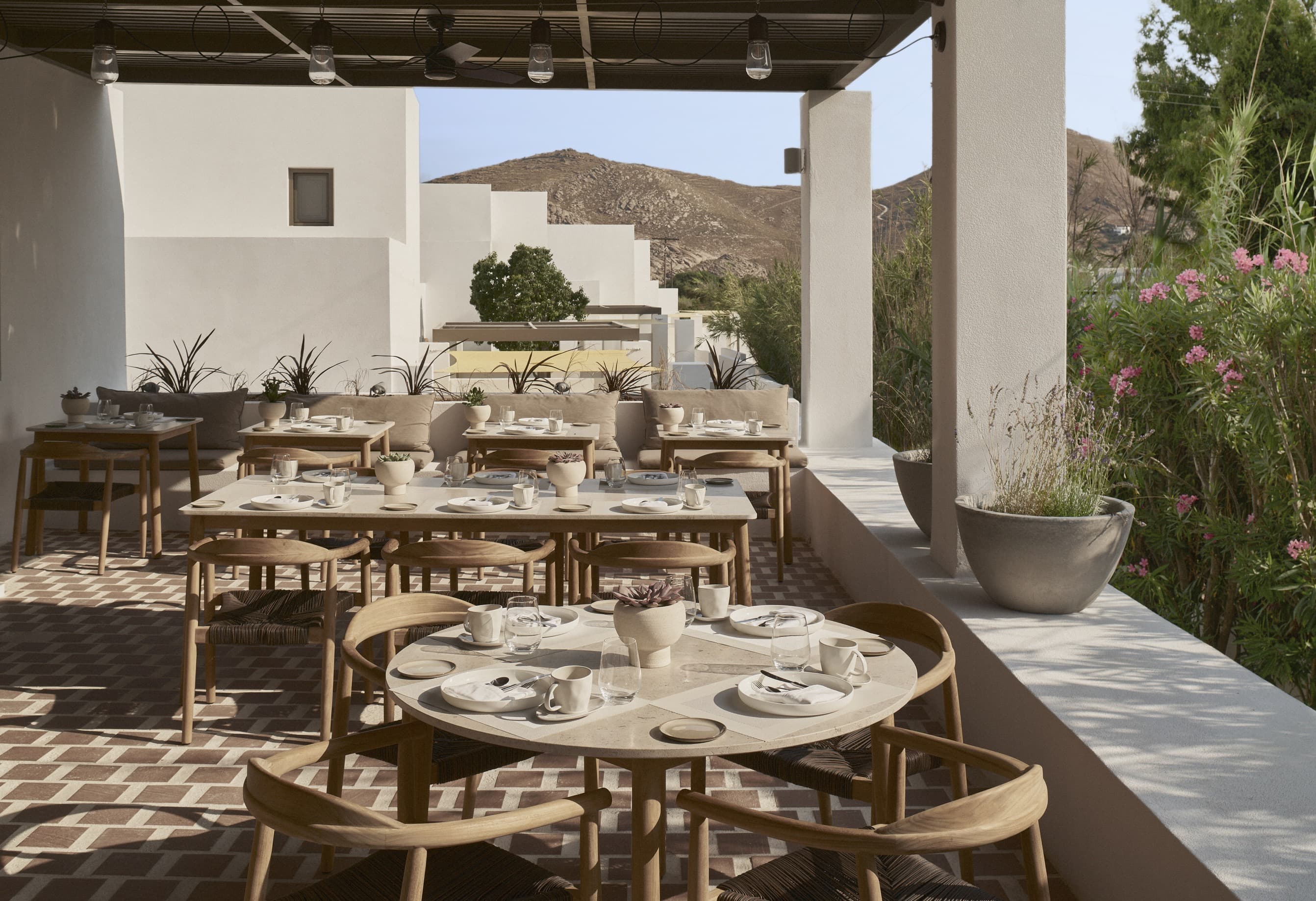 A sunlit terrace at Parīlio Hotel features round stone tables with wooden chairs, white tableware, and desert hills framed by minimalist architecture and potted flowers.
