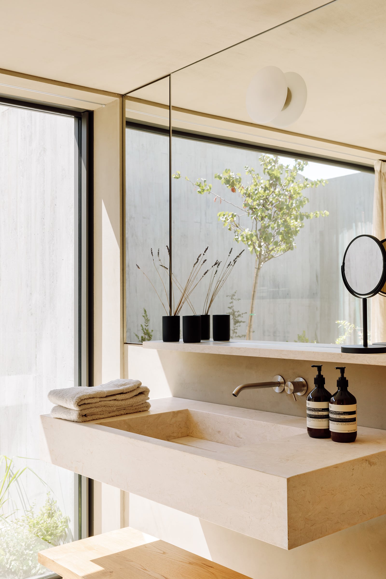Pa.te.os, Europe — Minimalist bathroom with limestone sink, brass fixtures, black ceramic planters, and floor-to-ceiling windows overlooking courtyard garden.