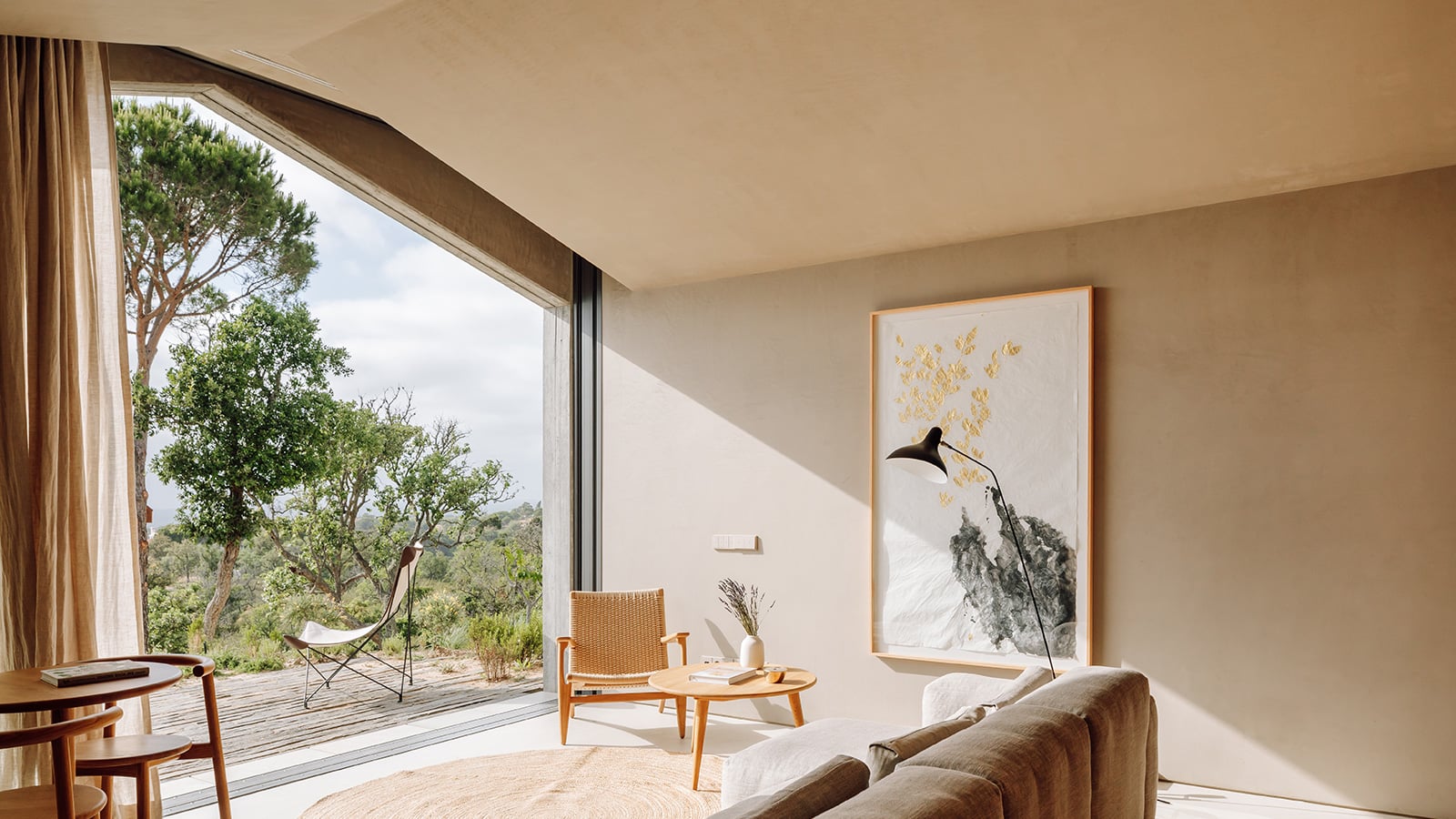 Pa.te.os, Europe — Minimalist living room with angled skylights, natural wood furnishings, black arc lamp, and panoramic forest views through floor-to-ceiling glazing.