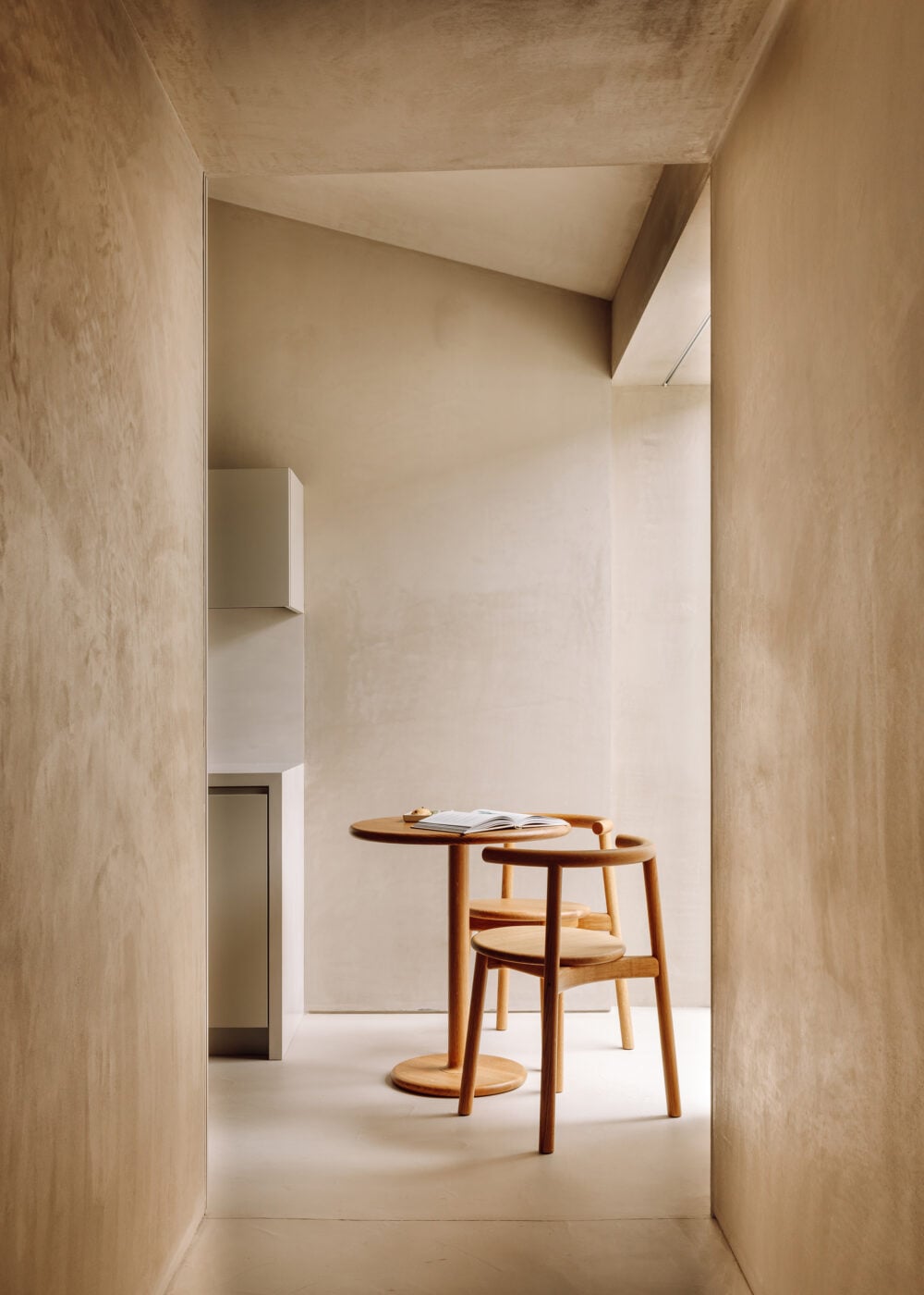 Pa.te.os, Europe — Minimalist stone corridor with wooden round table, chair, and skylight creating serene gallery space for reading and contemplation.