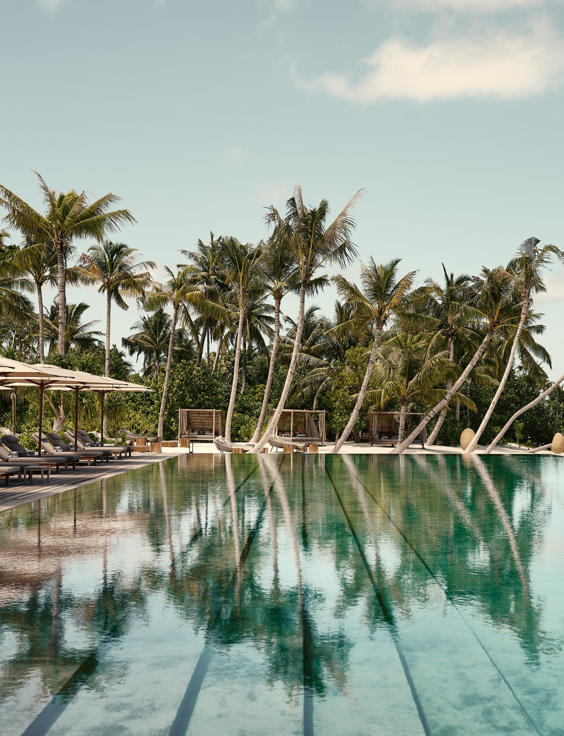 Patina Maldives, Asia — Infinity pool with palm reflections, teak deck, wooden cabanas, and turquoise water at tropical luxury resort.