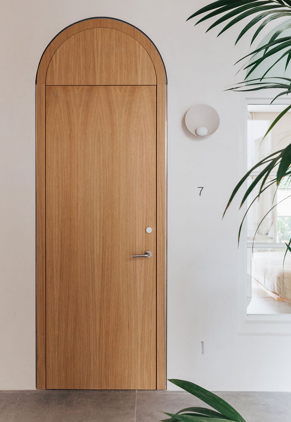 Plácido y Grata, Europe — Arched wooden door with horizontal grain detail and minimalist hardware, white walls with circular sconce, potted plant framing contemporary hospitality interior.