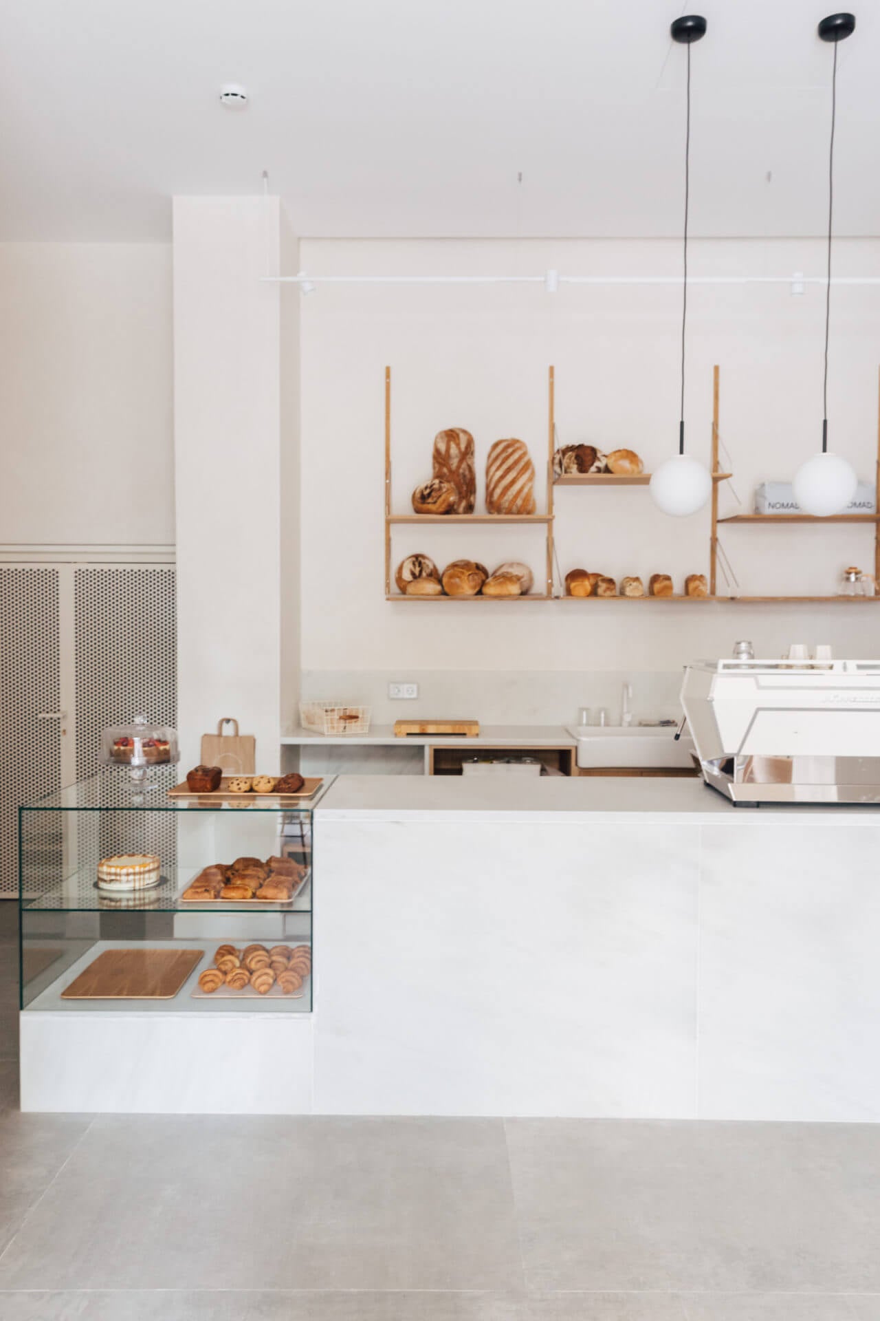 La Cafetería, Europe — Minimalist bakery counter with natural wood shelving, artisan breads, pendant lighting, and pristine white walls creating serene luxury hospitality design.