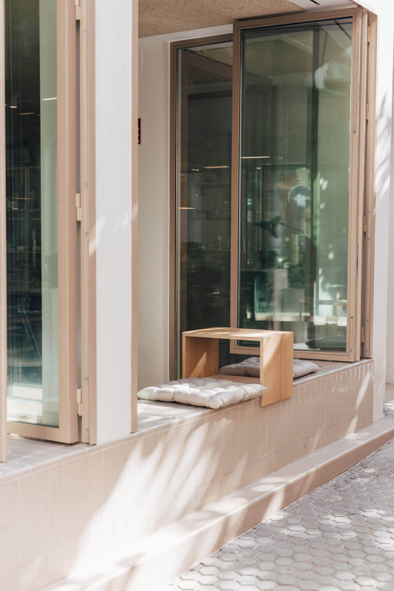 Plácido y Grata, Europe — Minimalist wooden window seat with cushions overlooking contemporary courtyard, natural light filtering through glass doors.
