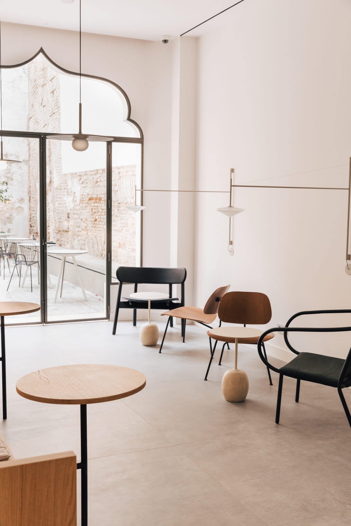 La Coctelería, Europe — Minimalist hospitality lounge with natural wood tables, black metal chairs, cream walls, and modernist pendant lighting overlooking courtyard.