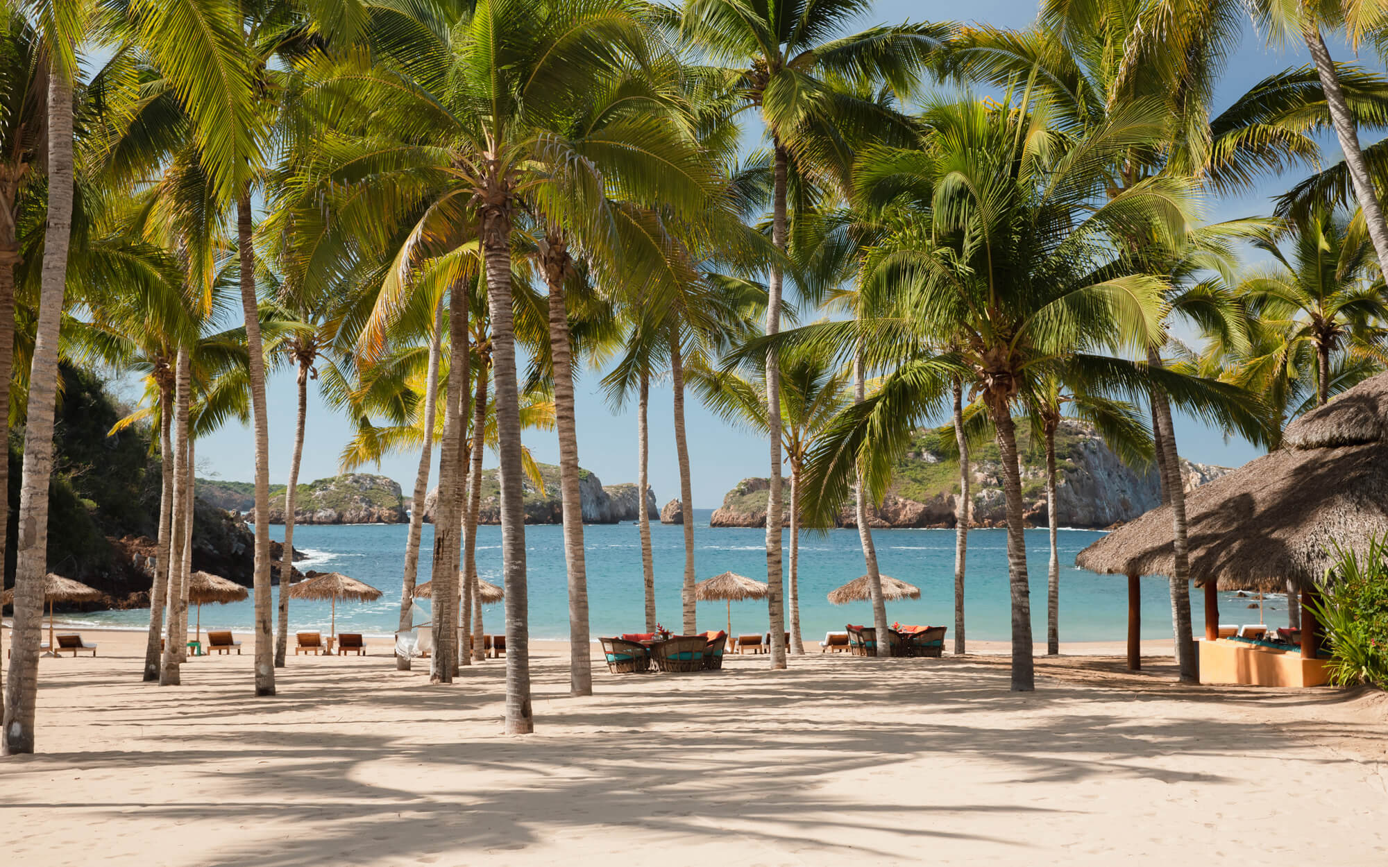 Caleta Blanca, Mexico — Palm-lined beach with thatched palapas, turquoise water, and limestone cliffs under clear sky.