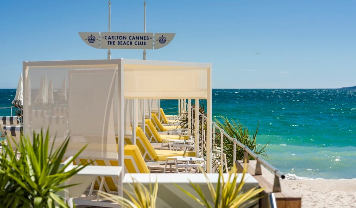 Carlton Beach Club — Carlton Cannes The Beach Club, France — Luxury cabanas with yellow loungers overlooking turquoise Mediterranean sea, minimalist white architecture and striped awnings.