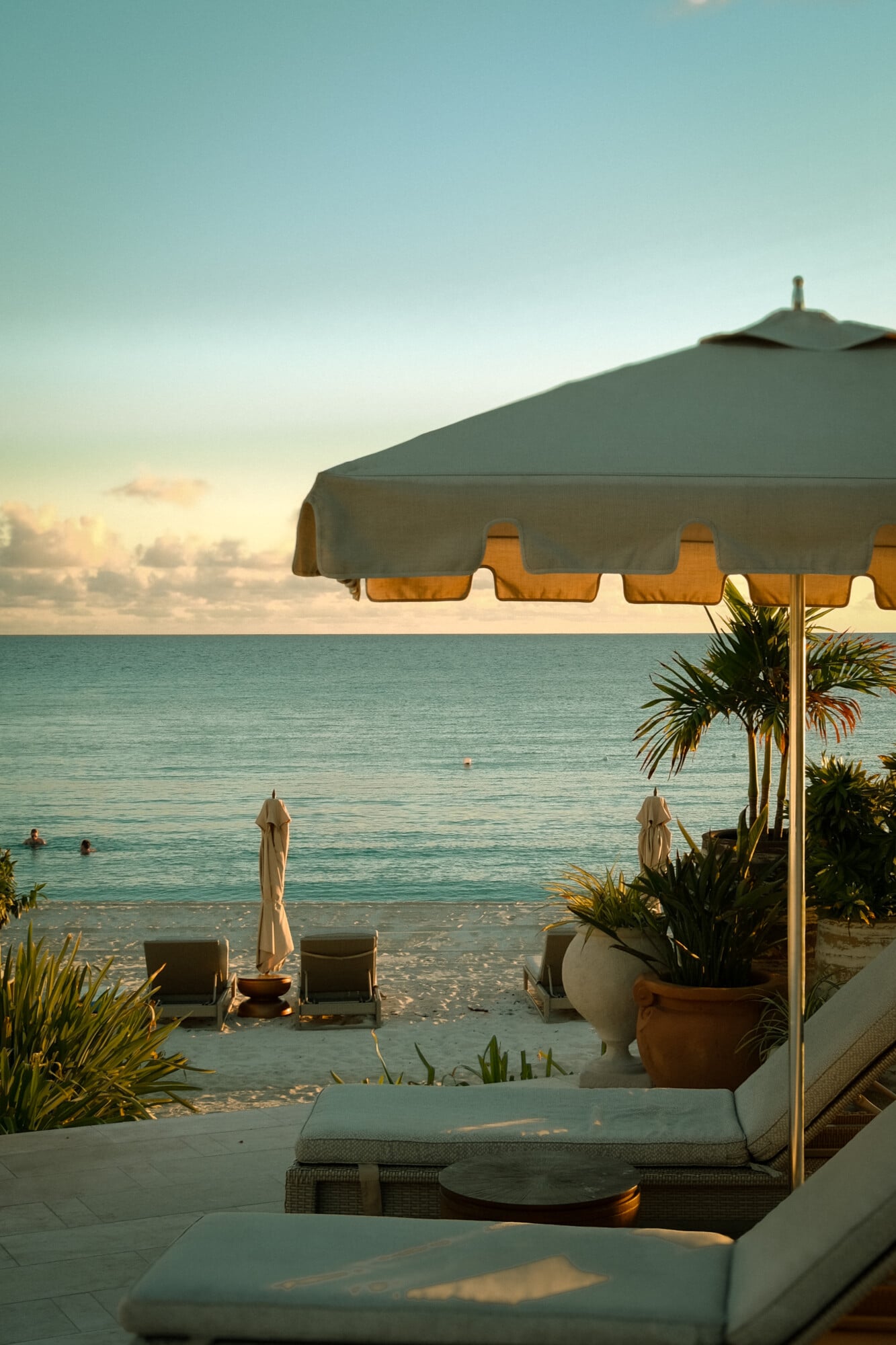 Cap Juluca, Anguilla — Gray striped beach umbrella with golden scalloped trim overlooking turquoise Caribbean waters at sunset, lounge seating and tropical plantings visible.