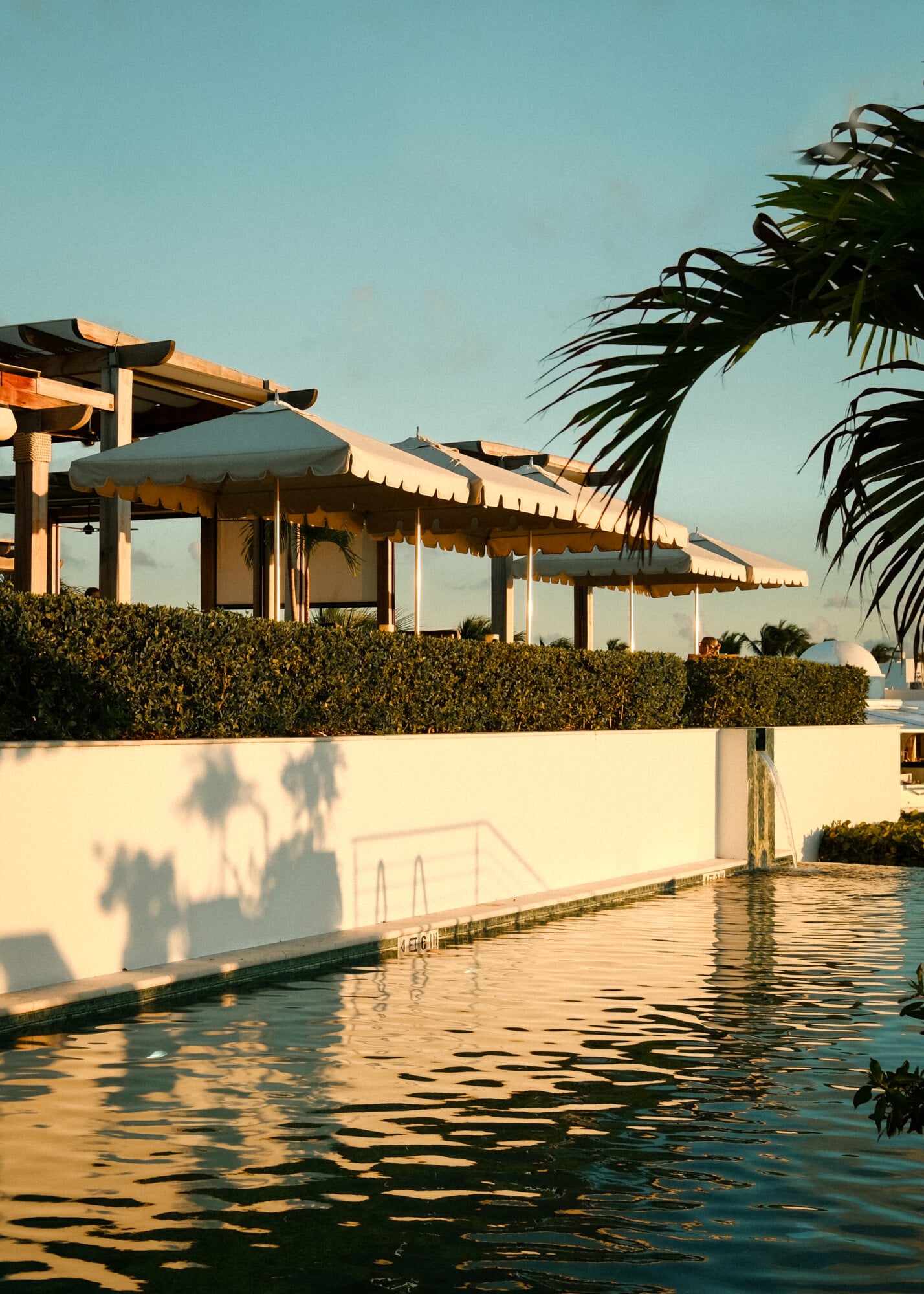 Cap Juluca, Anguilla — Modernist poolside pavilions with cream awnings at sunset, palm fronds casting shadows on whitewashed walls, turquoise water reflecting golden hour light.