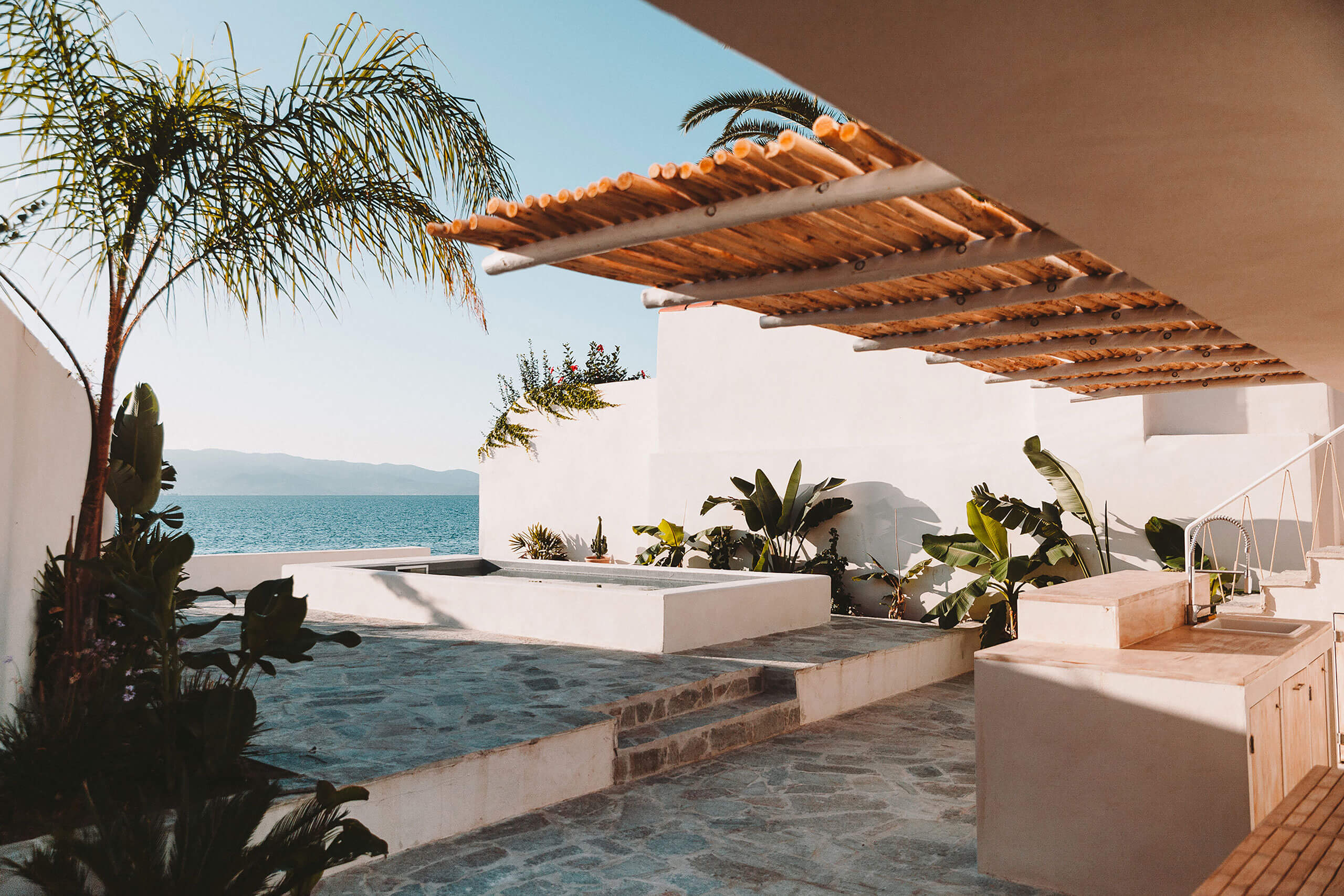 Casa Santa Teresa, Europe — Whitewashed Mediterranean villa terrace with wooden pergola, sea views, tropical plants, and stone steps at golden hour.