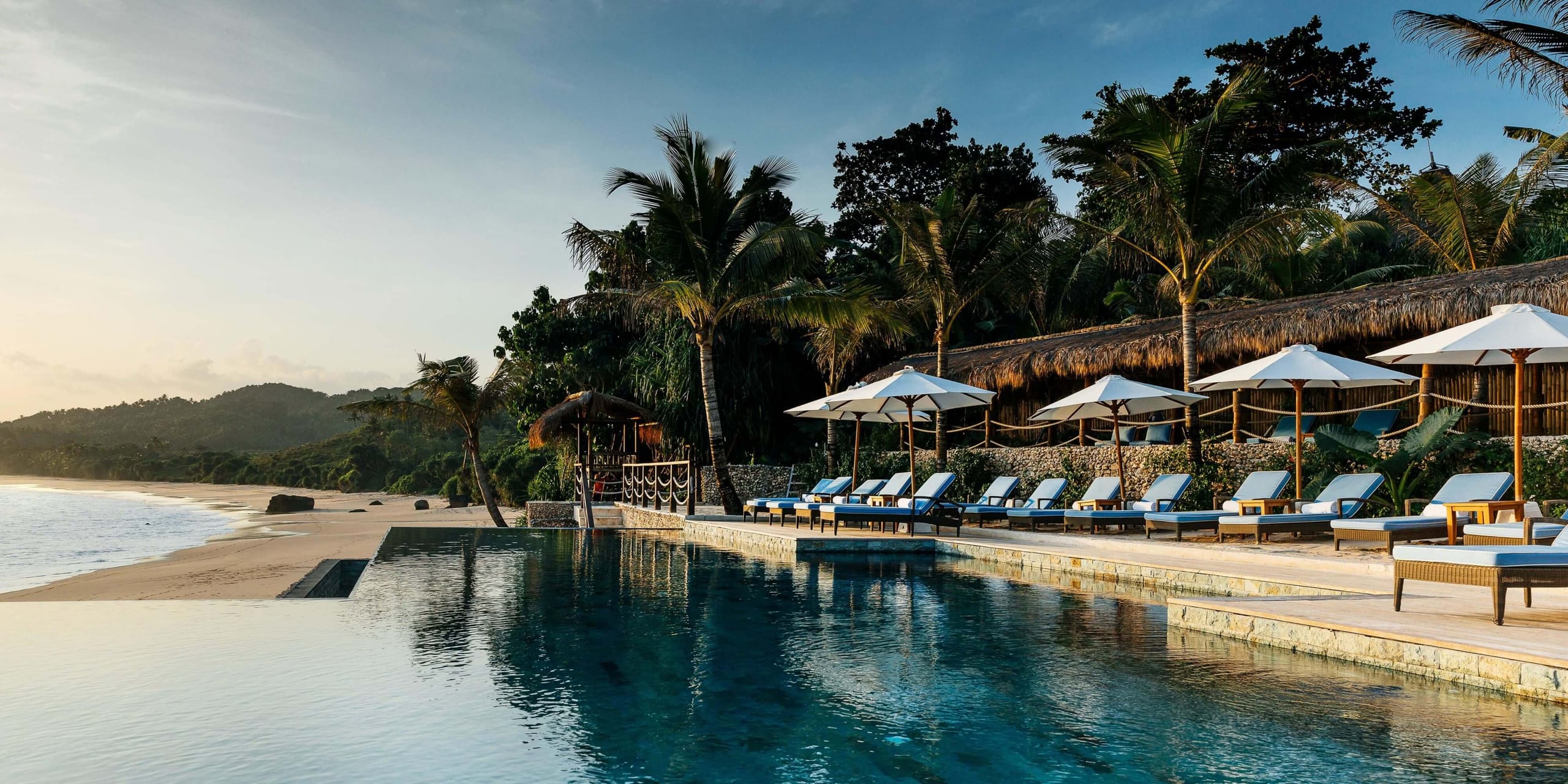 NIHI Sumba, Indonesia — Infinity pool with teak loungers and white umbrellas overlooking pristine beach, palm trees, and forested hillside at sunset.