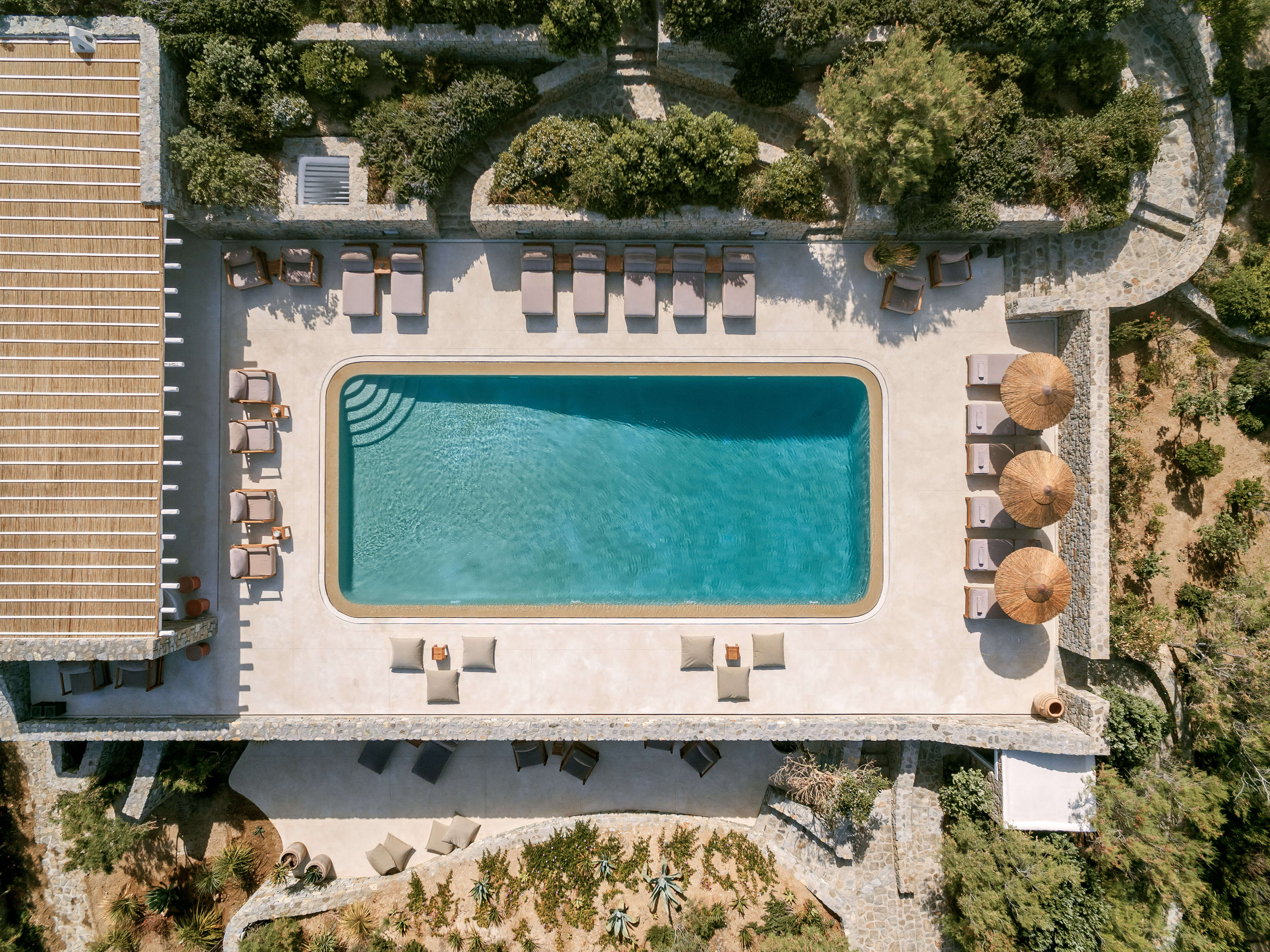 Villa Tama, Europe — Aerial view of modernist pool deck with turquoise rectangular pool, lounge chairs, woven umbrellas, and manicured gardens surrounding pale stone terrace.
